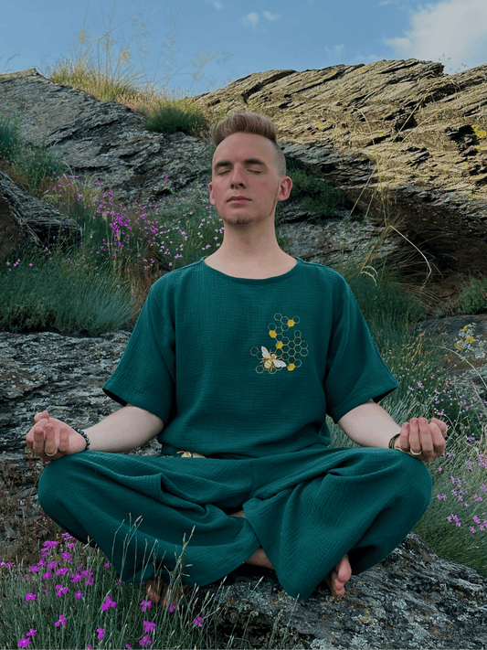 Person in a green outfit meditating on a rocky hillside with flowers and sky.