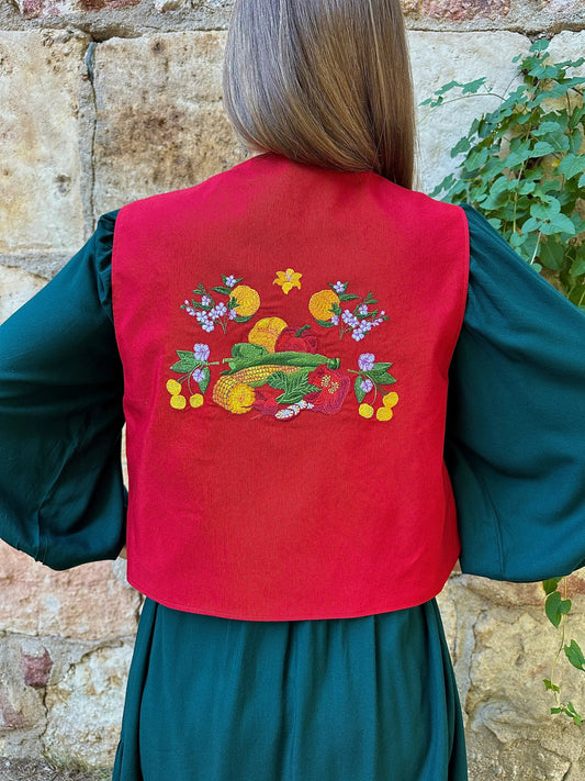 Person wearing a red vest with embroidered vegetables and flowers, standing against a stone wall.
