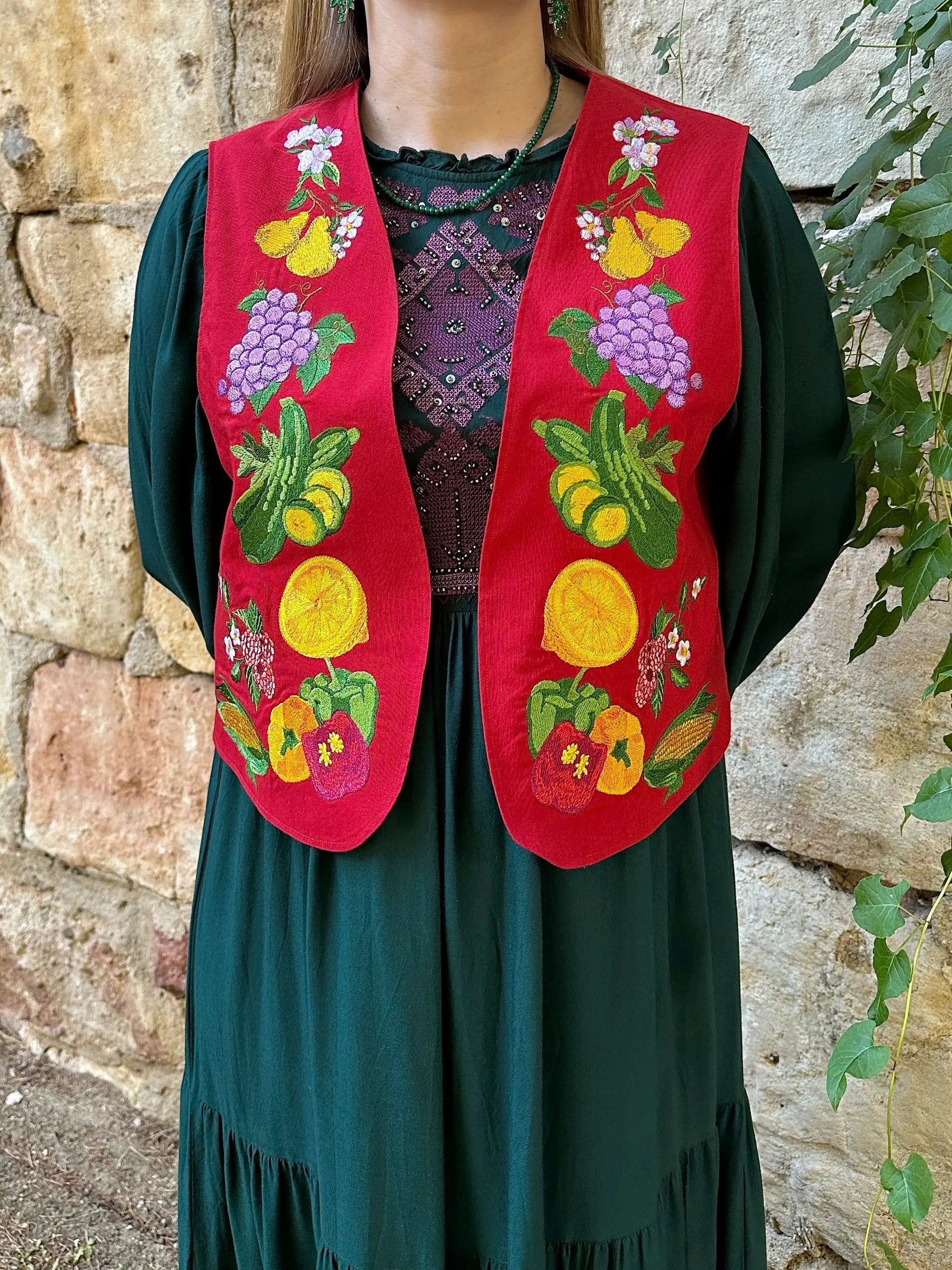Person wearing a green dress with a red embroidered vest featuring fruits and flowers against a stone wall.