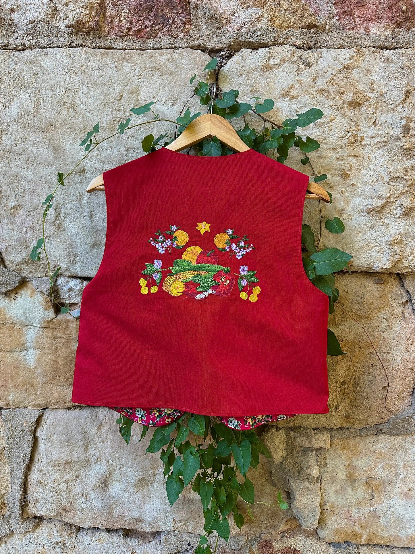 Red embroidered top on a hanger against a stone wall with greenery.