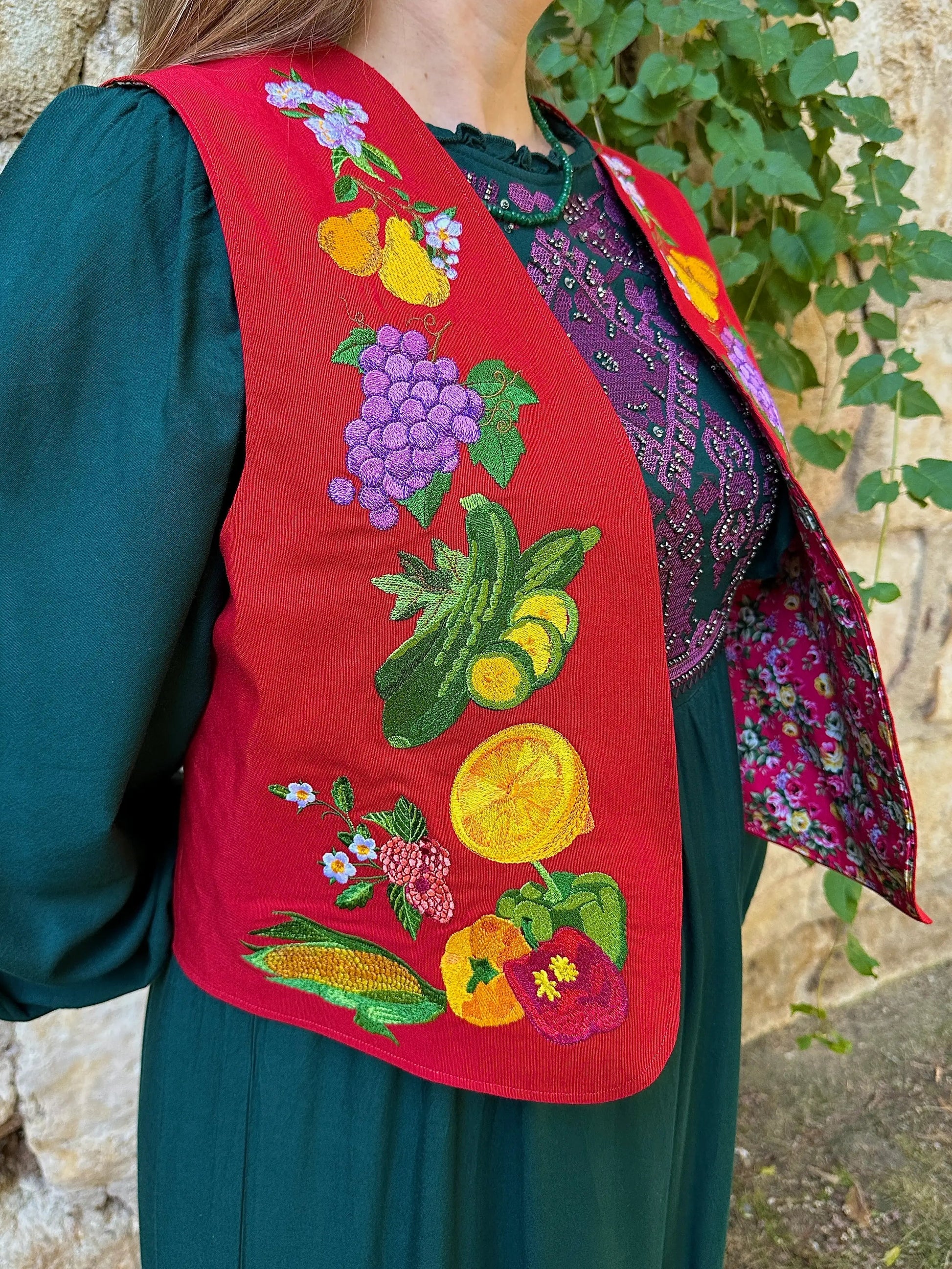 Person wearing a red embroidered vest with colorful vegetable patterns, standing against a stone wall.