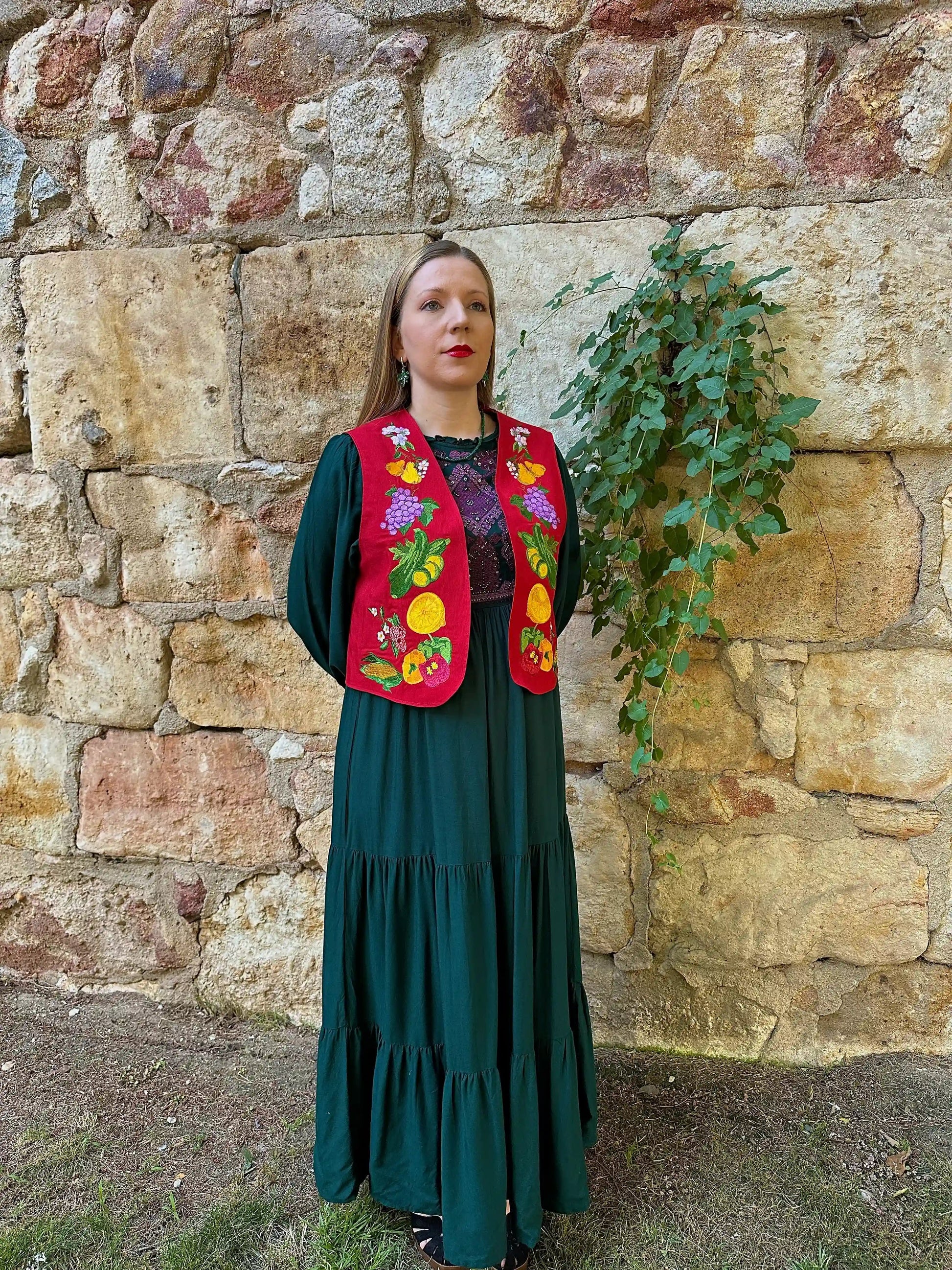 Woman wearing a green dress and colorful embroidered vest against a stone wall.