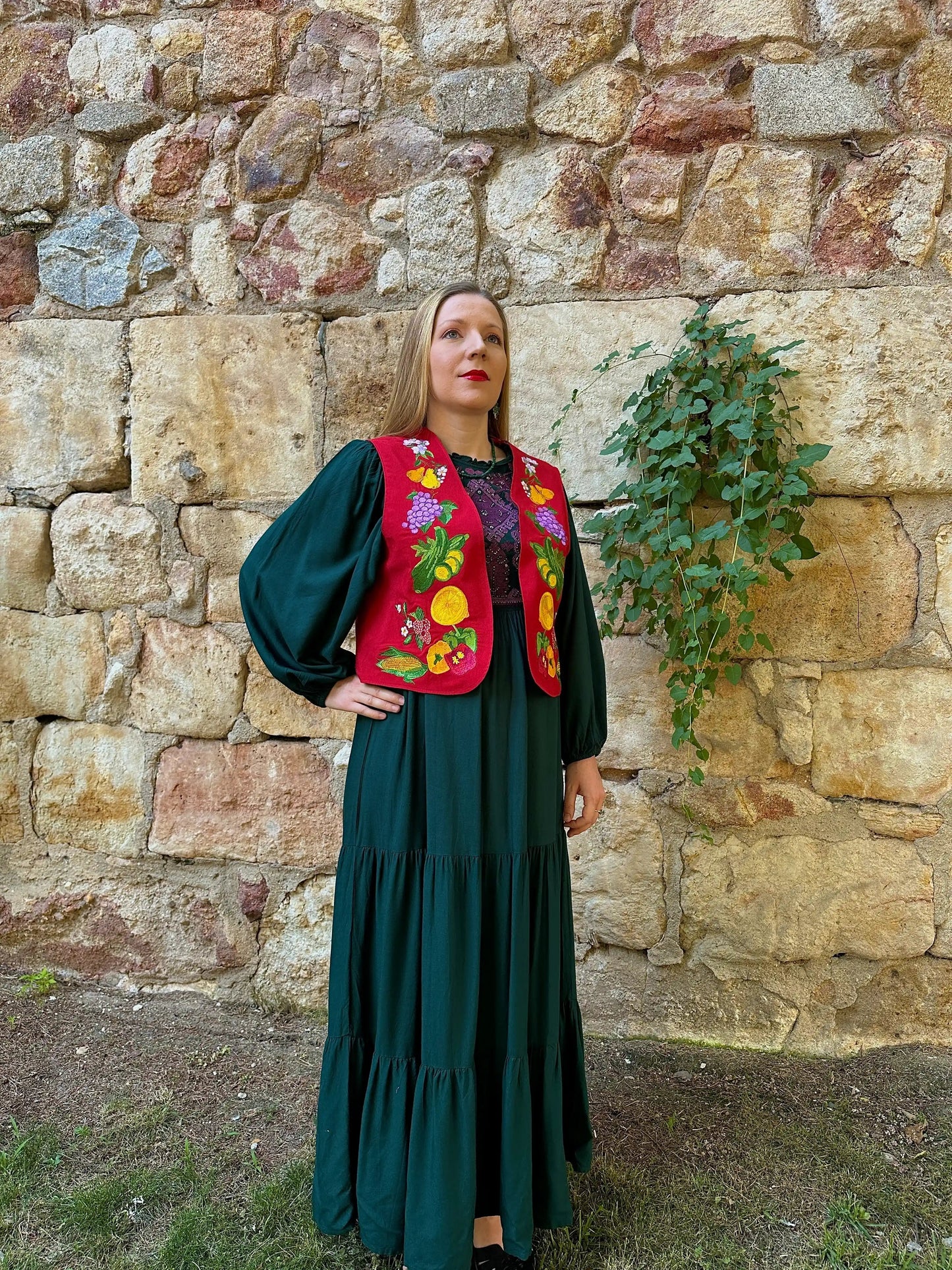Woman wearing a dark green dress and colorful embroidered vest against a stone wall.