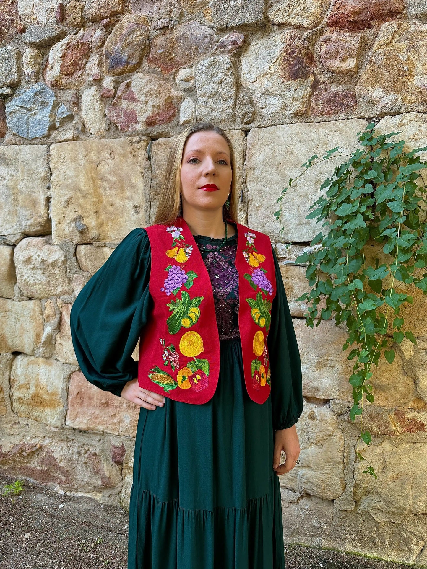Woman wearing a colorful embroidered vest against a stone wall.
