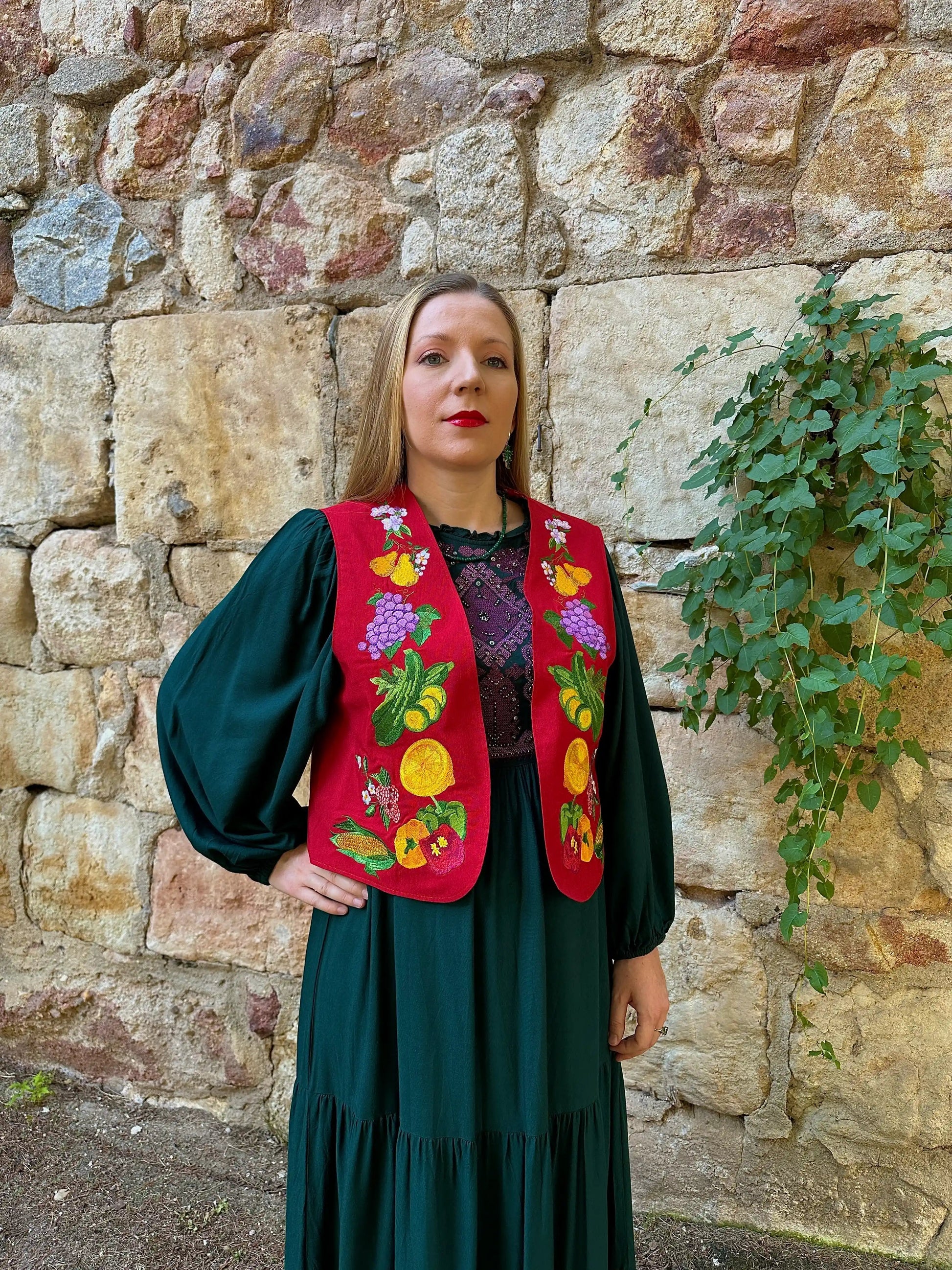 Woman wearing a colorful embroidered vest against a stone wall.