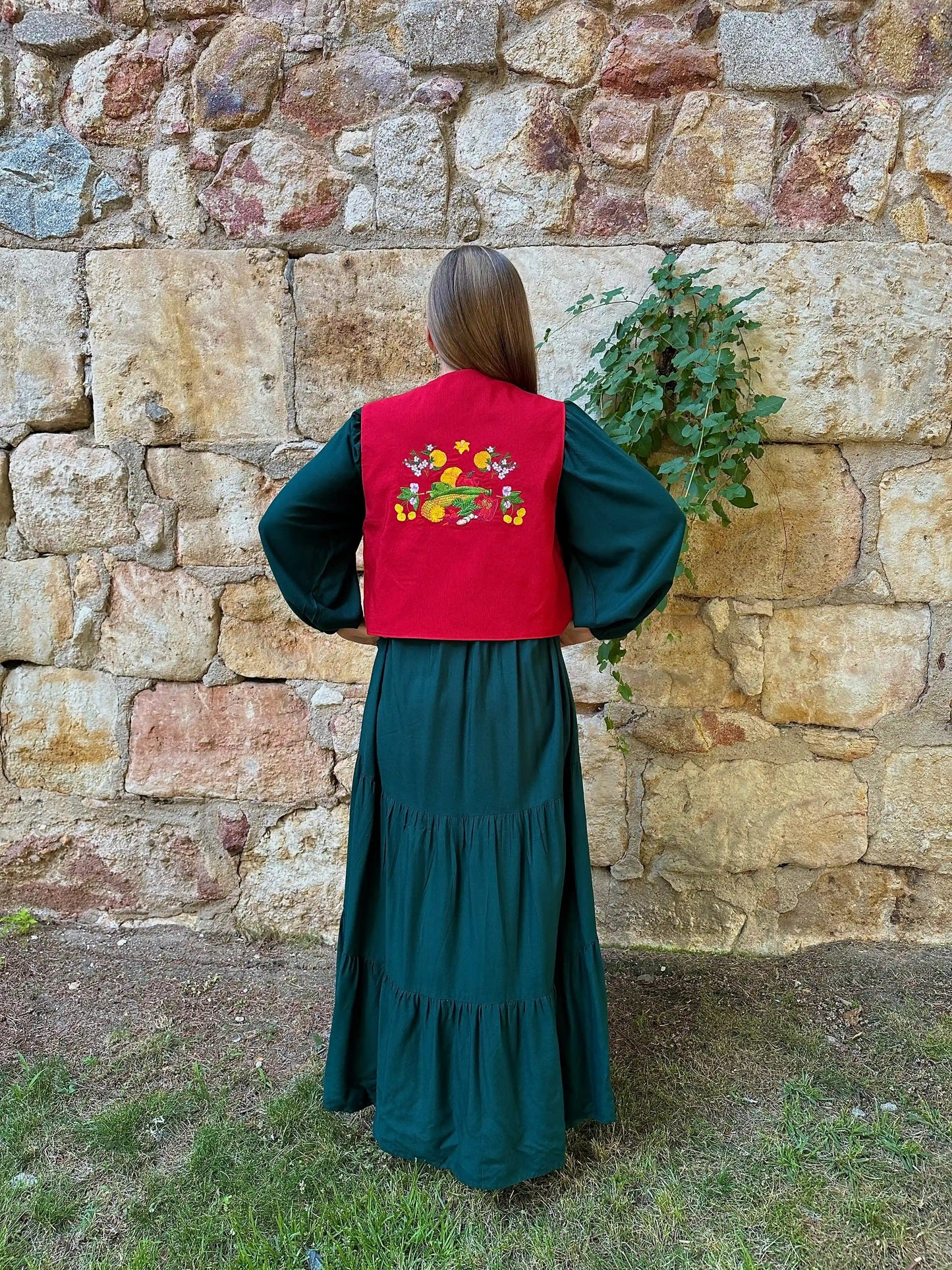 Person wearing a green dress and red vest with floral embroidery against a stone wall.