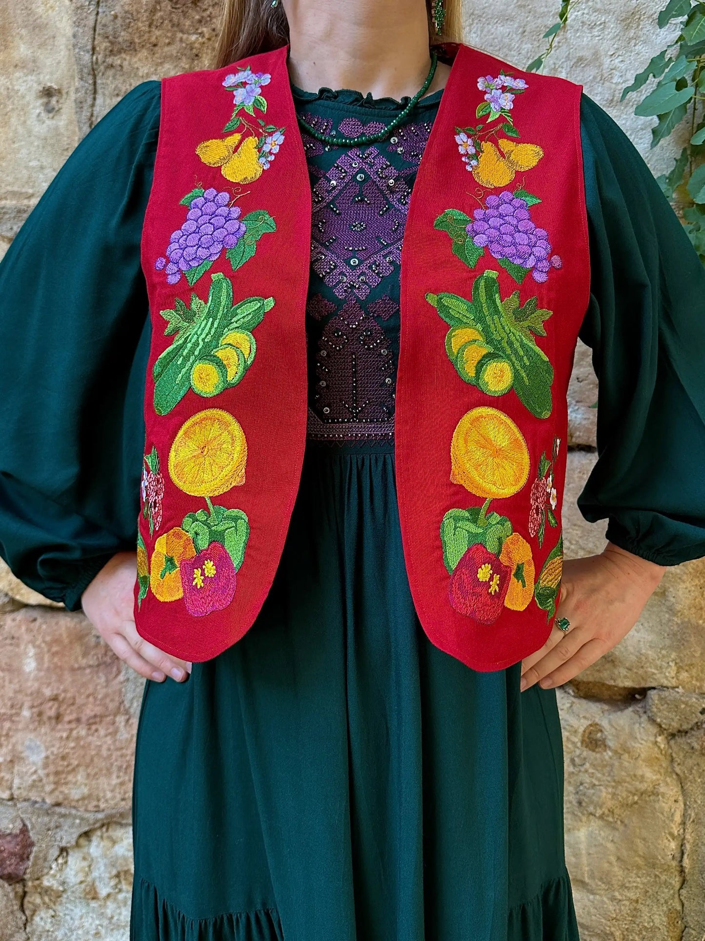 Person wearing a red embroidered vest with fruit designs against a stone wall.