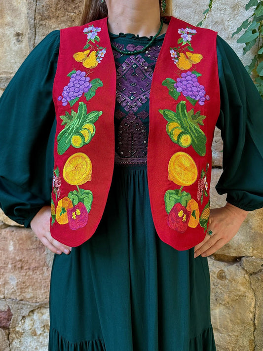 Person wearing a red embroidered vest with fruit designs against a stone wall.