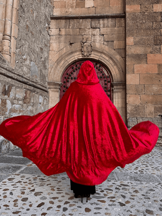 Person wearing a red hooded cloak standing in front of a stone building.