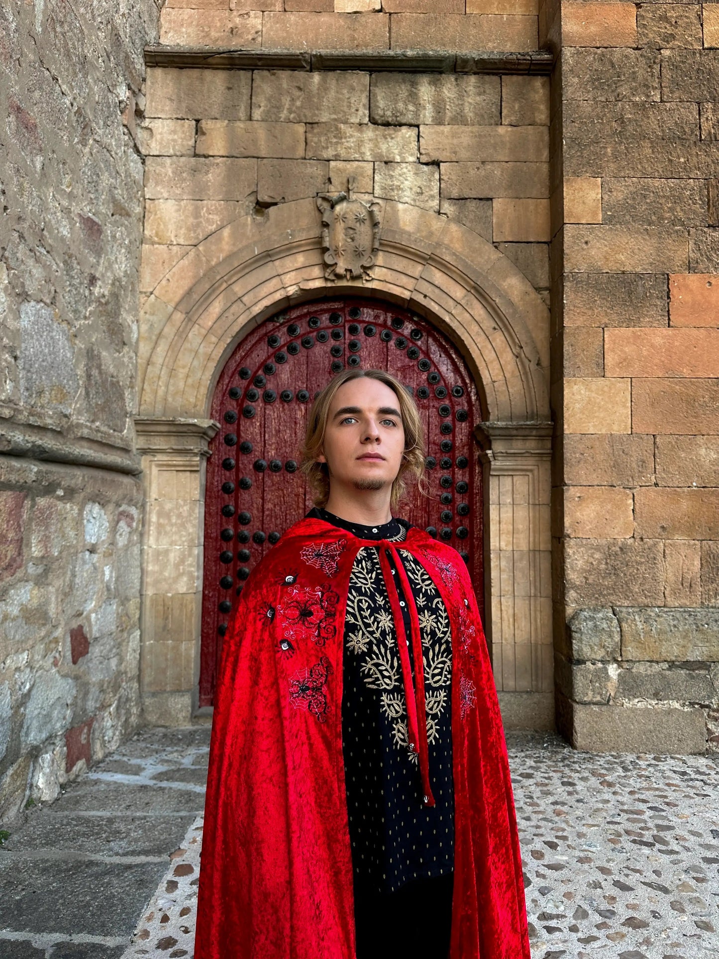 Person wearing a red embroidered cape in front of a stone archway.