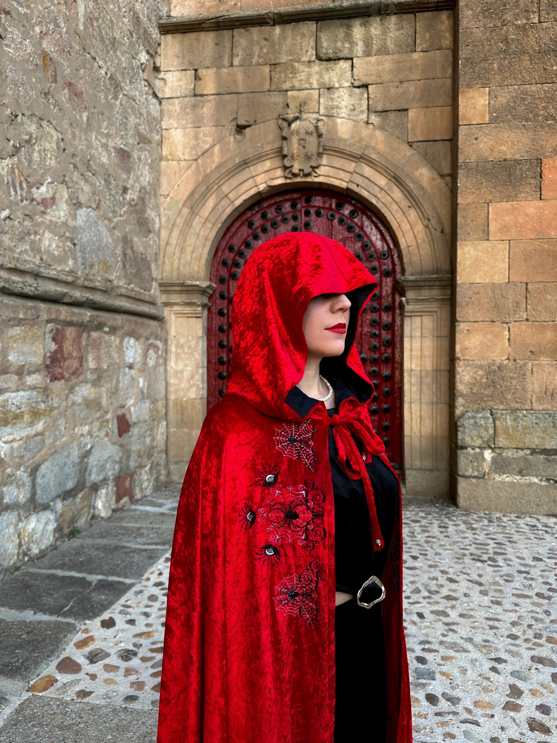 Person wearing a red hooded cloak with black embroidery standing in front of a stone building.