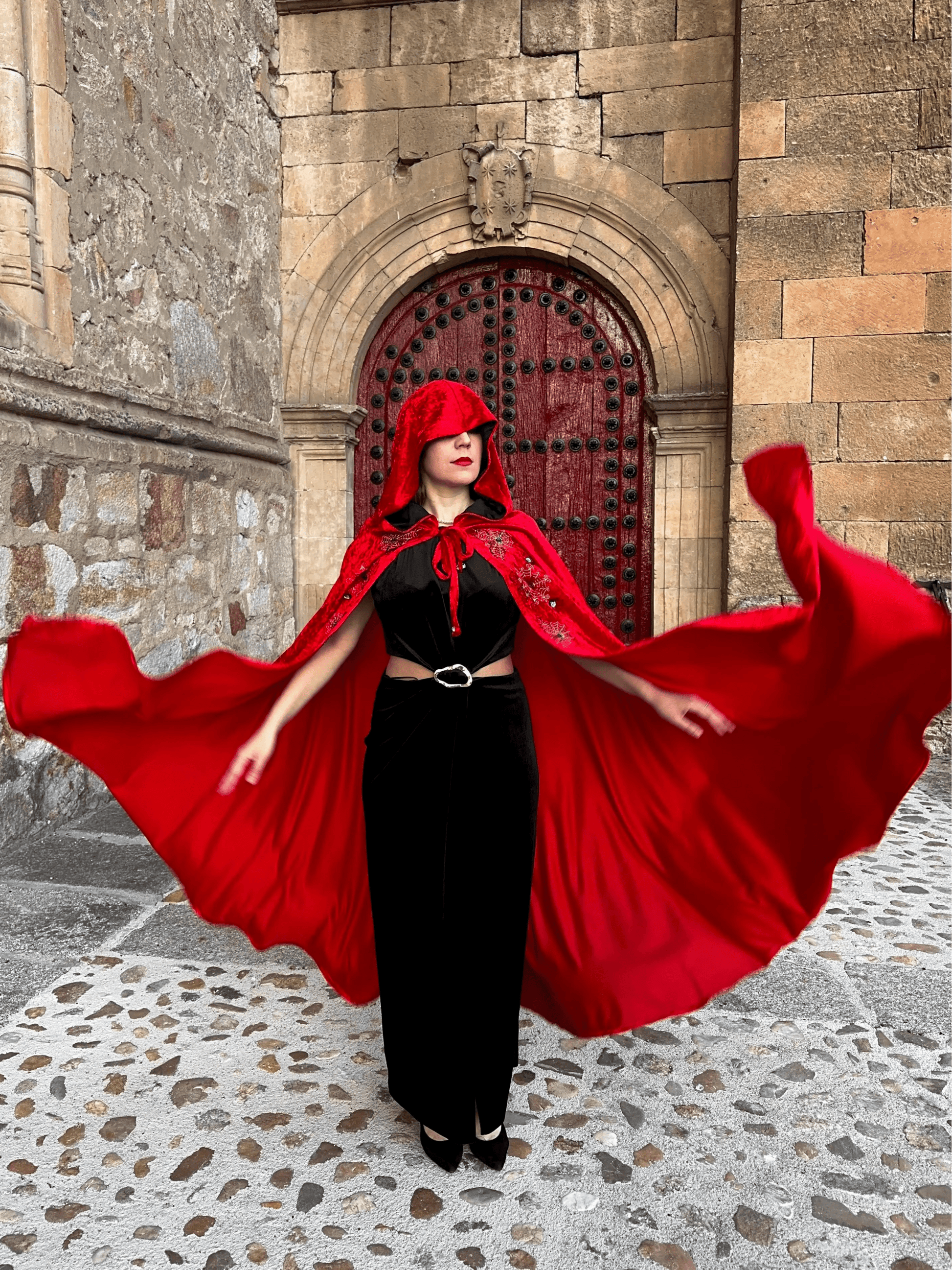 Person wearing a red cloak and black dress in front of a stone building with a red door.