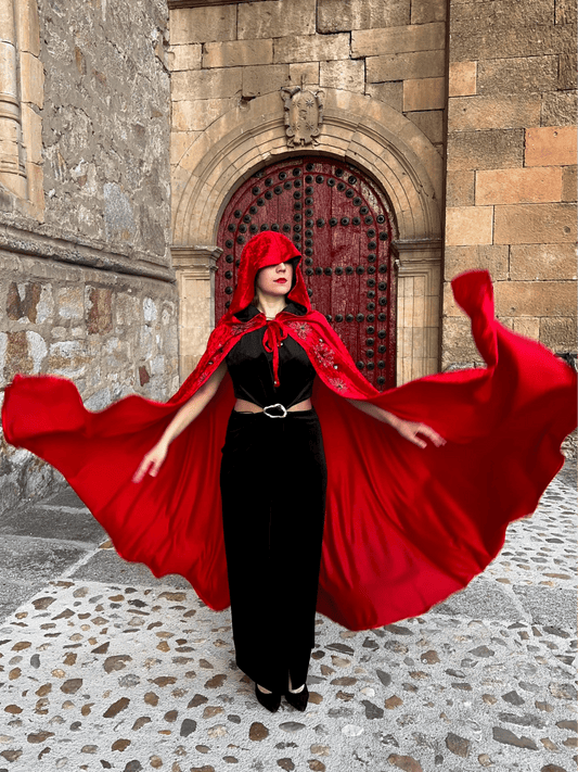 Person wearing a red cloak and black dress in front of a stone building with a red door.