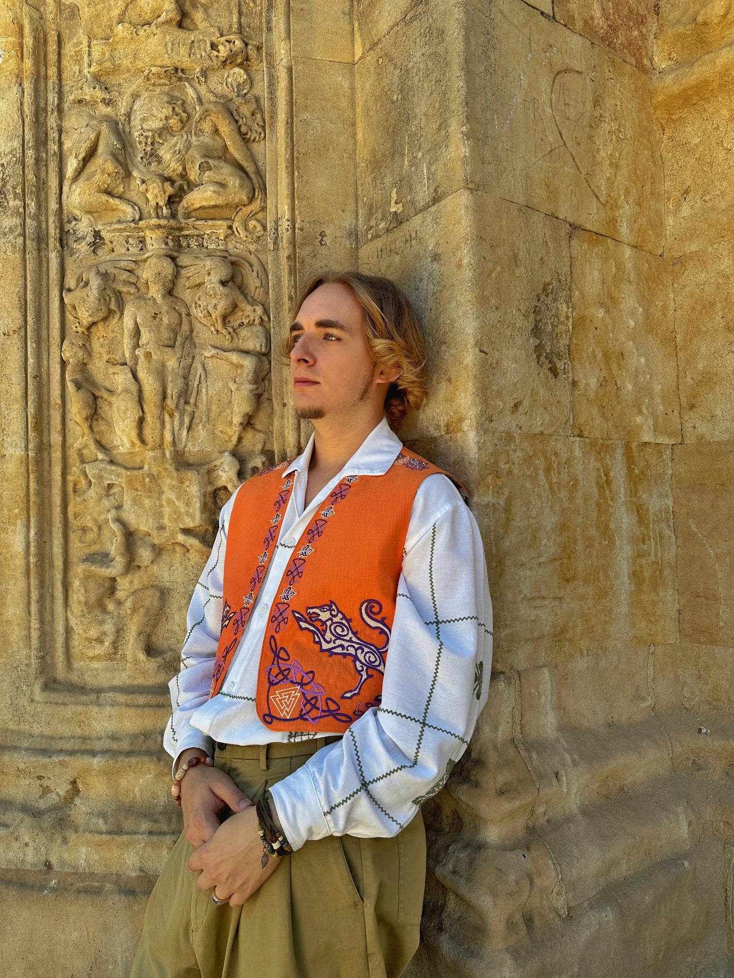 Person wearing an orange vest with embroidery against a stone wall with carvings
