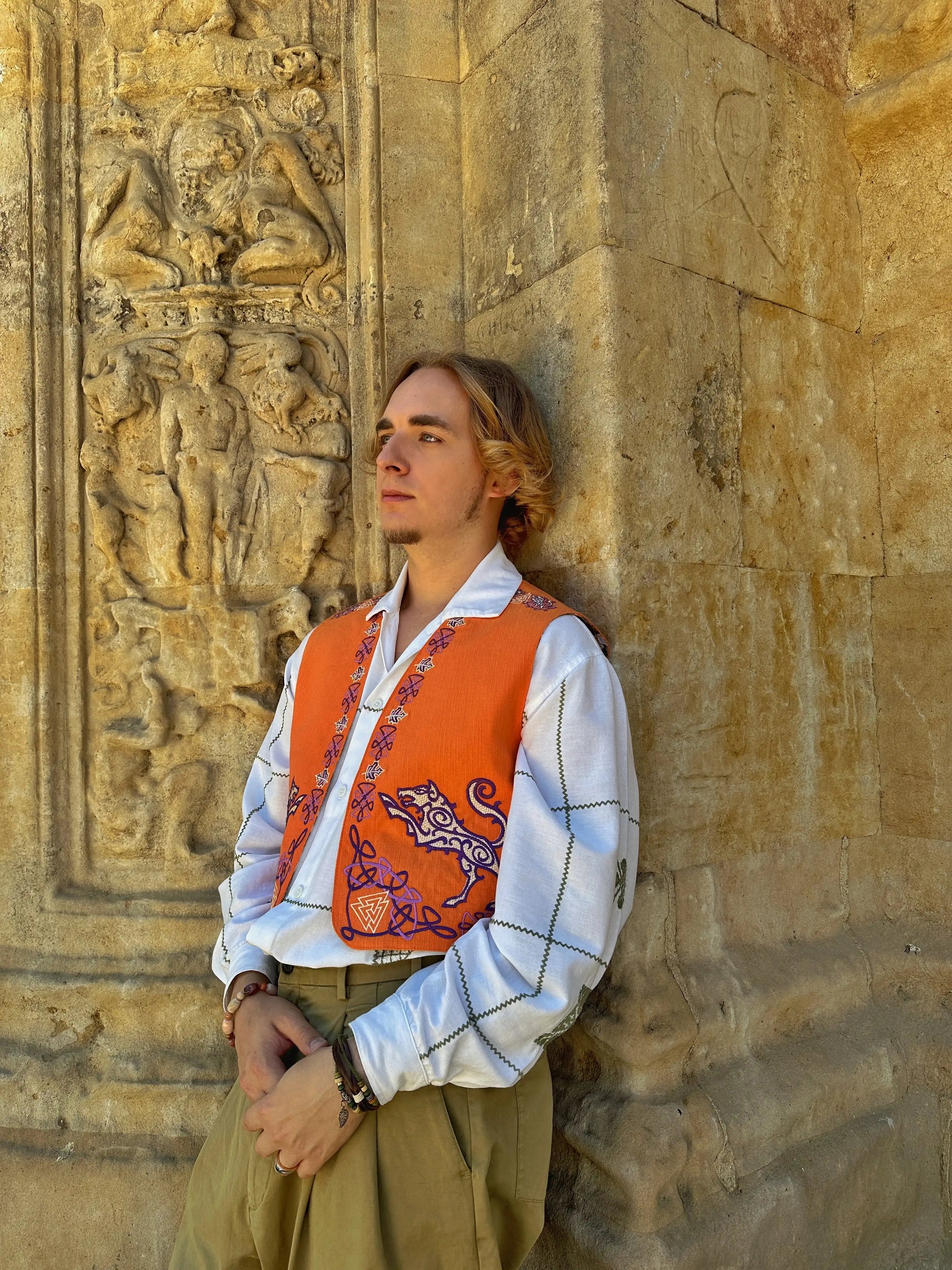 Person wearing an orange vest with embroidery against a stone wall with carvings
