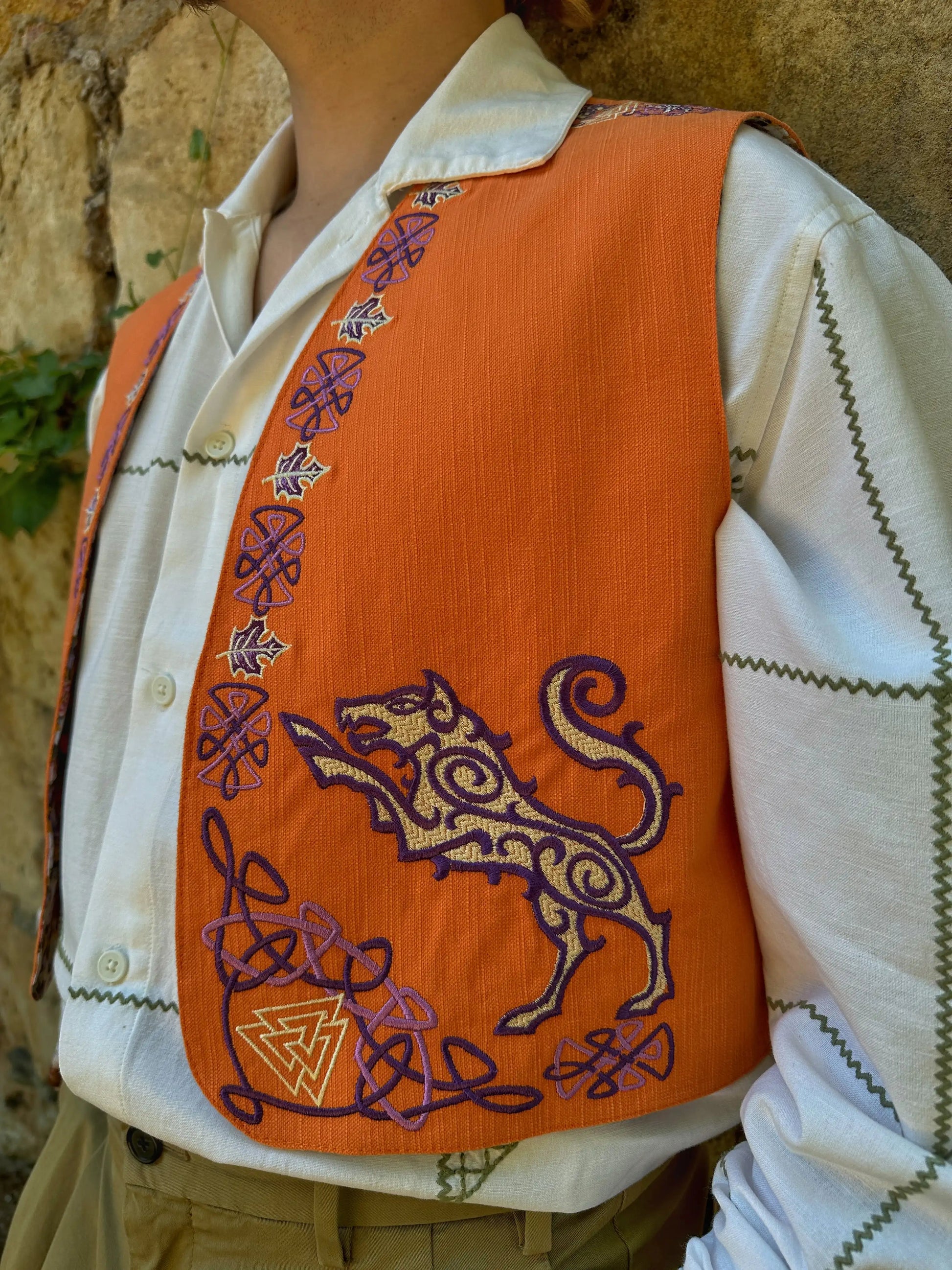 Person wearing an orange embroidered vest with a lion design against a stone wall.