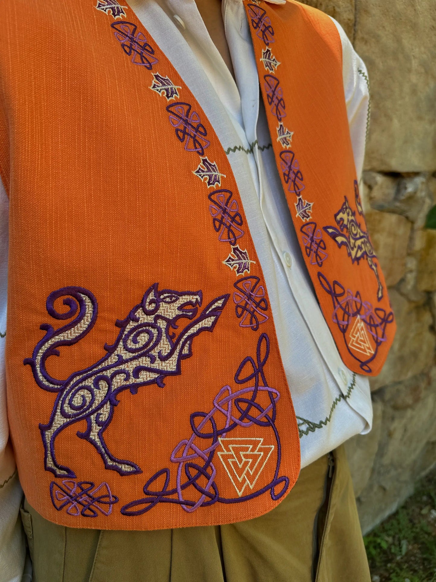 Orange embroidered vest with a purple lion design against a stone wall background