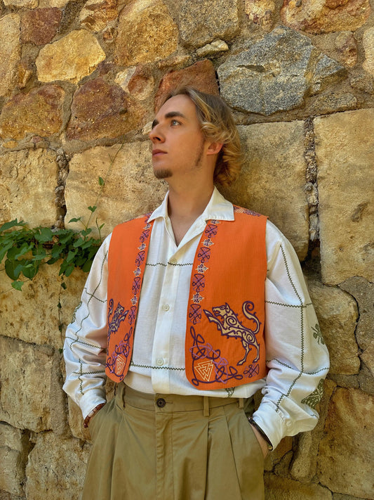 Man wearing an orange embroidered vest over a white shirt against a stone wall.