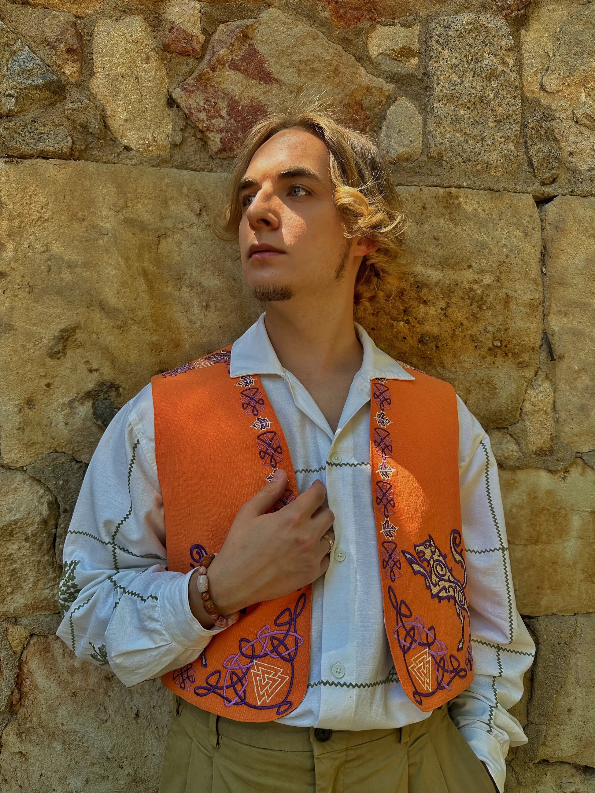 Man wearing an orange embroidered vest against a stone wall