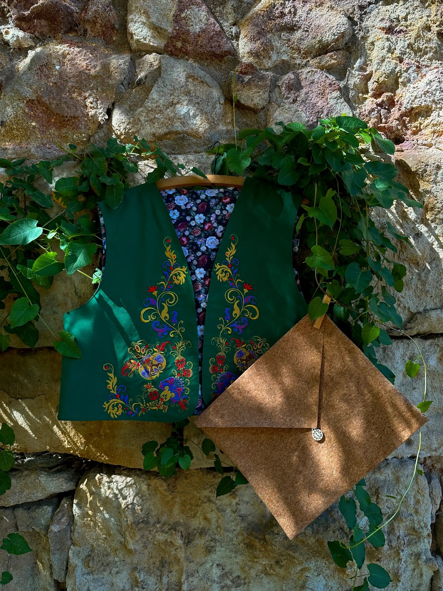 Green embroidered vest and brown bag hanging on a stone wall with greenery.