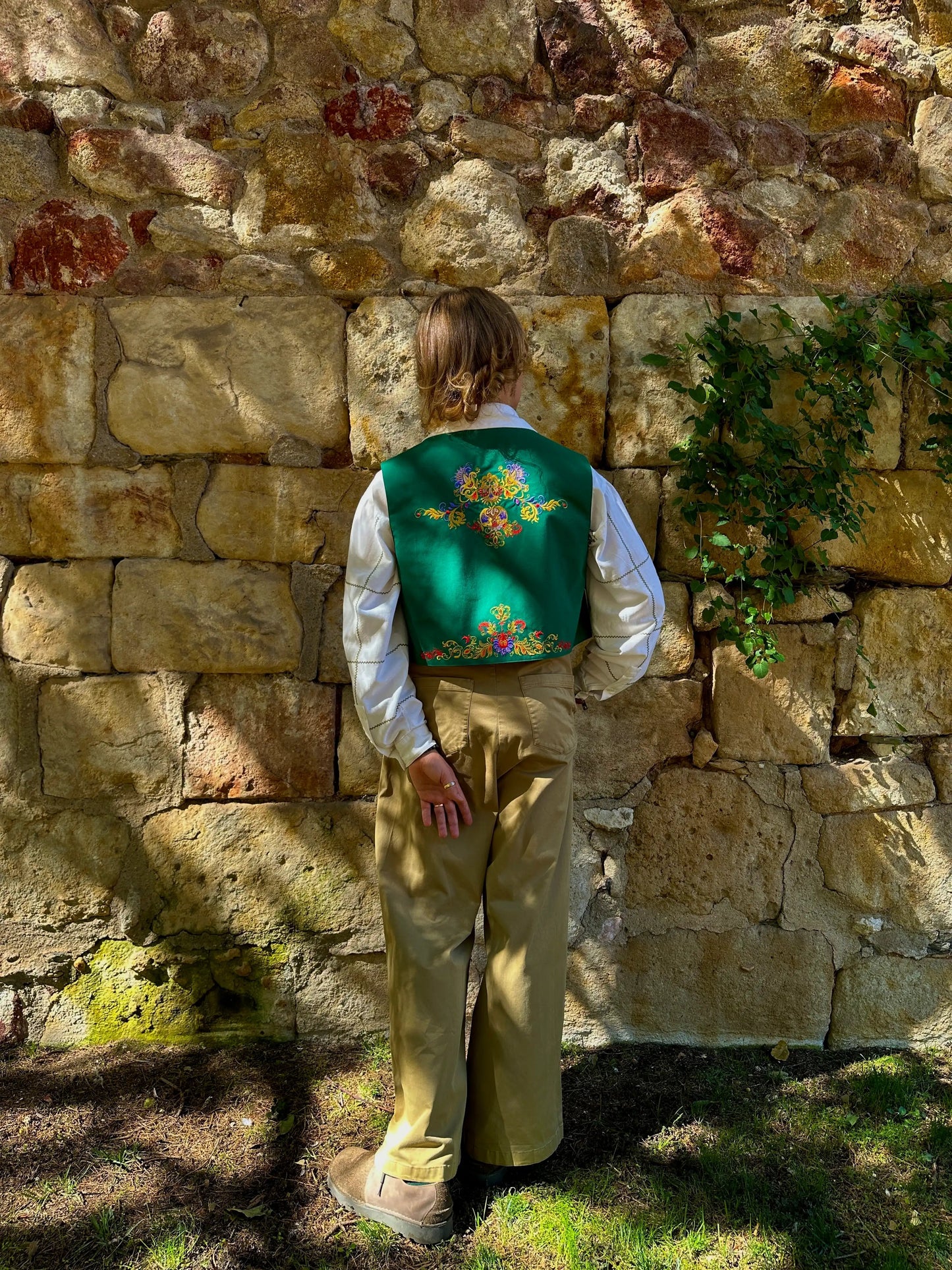 Person wearing a traditional outfit with a green vest against a stone wall.