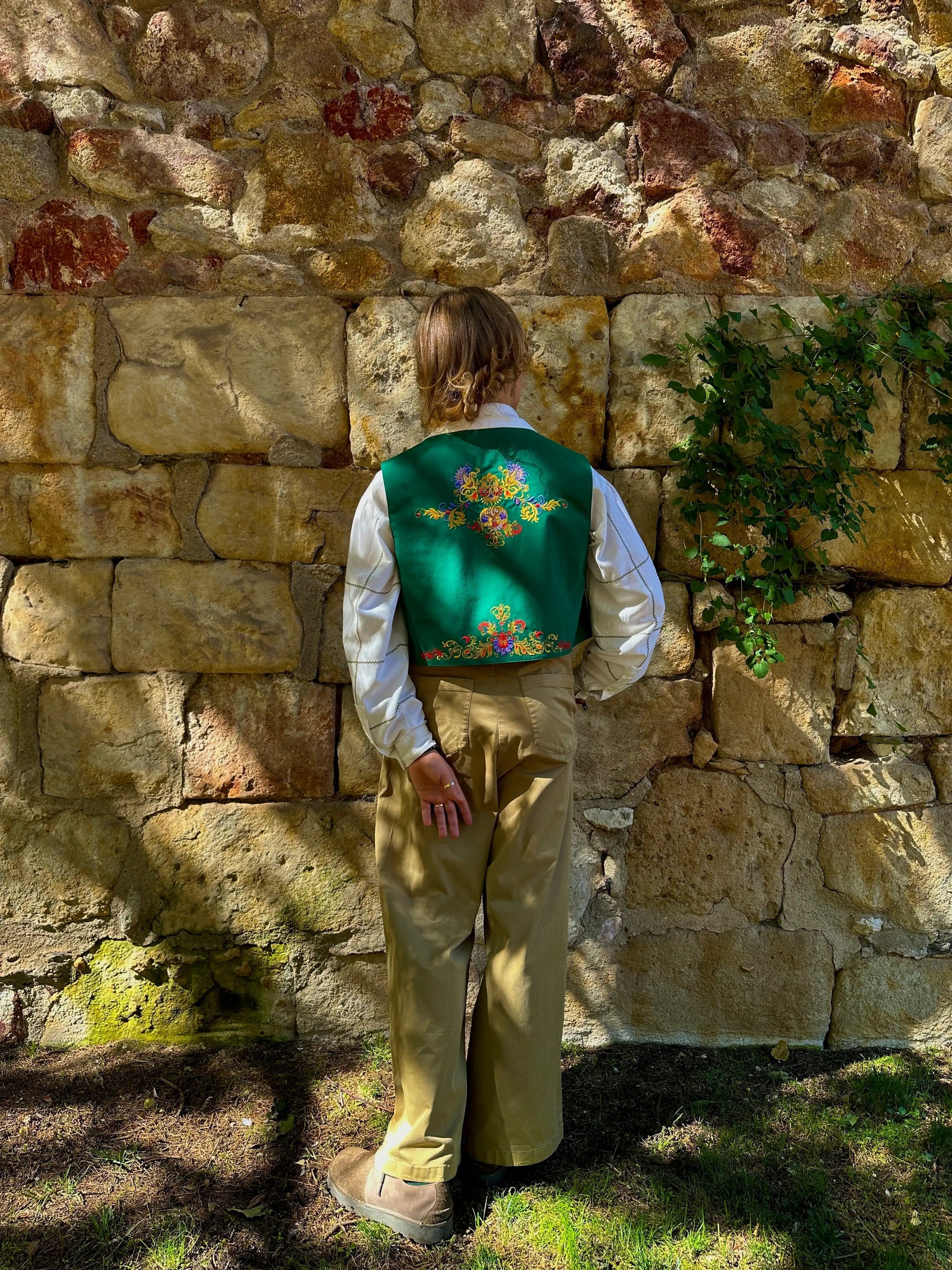 Person wearing a traditional outfit with a green vest against a stone wall.