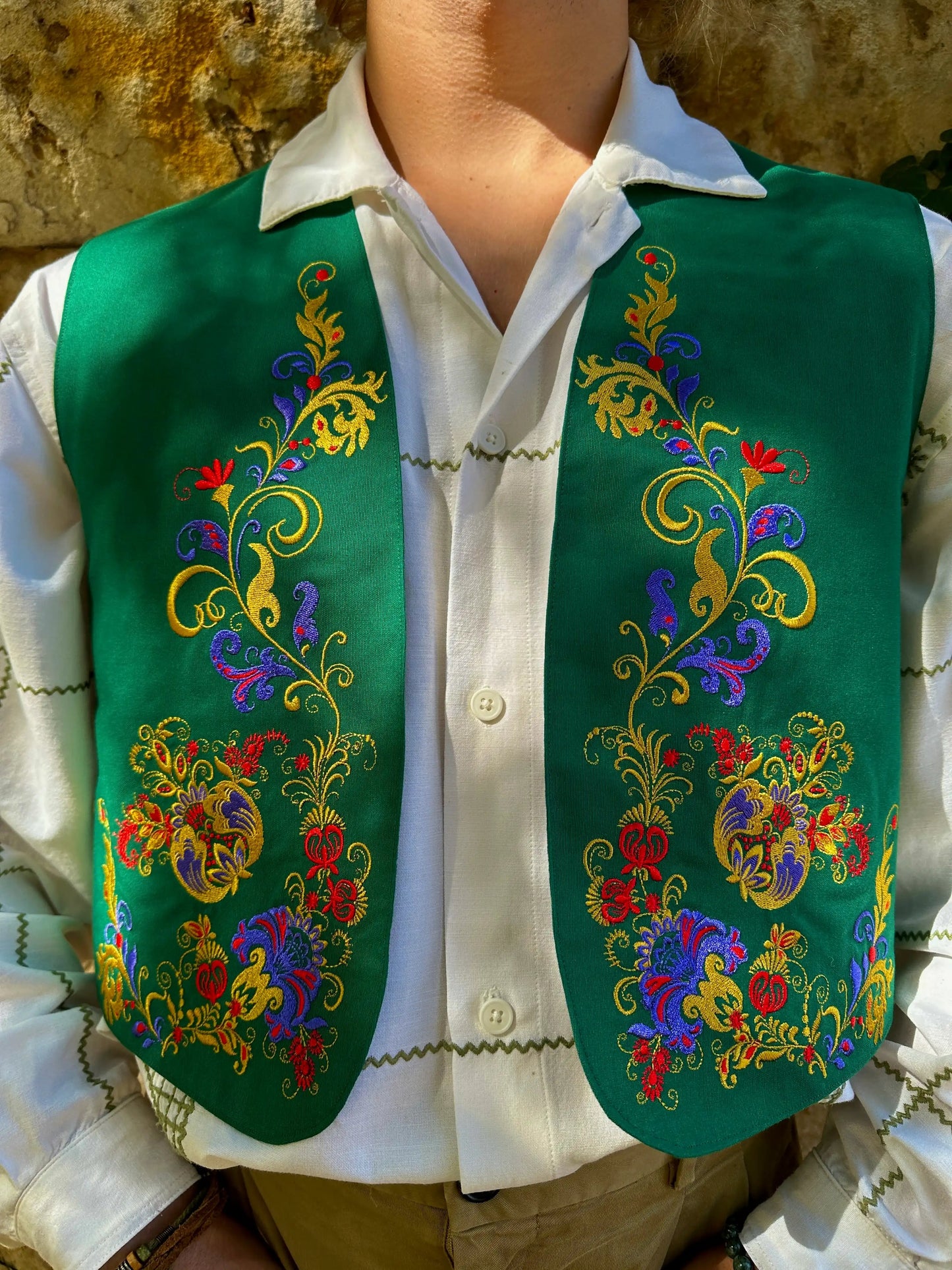 Person wearing a green embroidered vest over a white shirt with a stone wall background