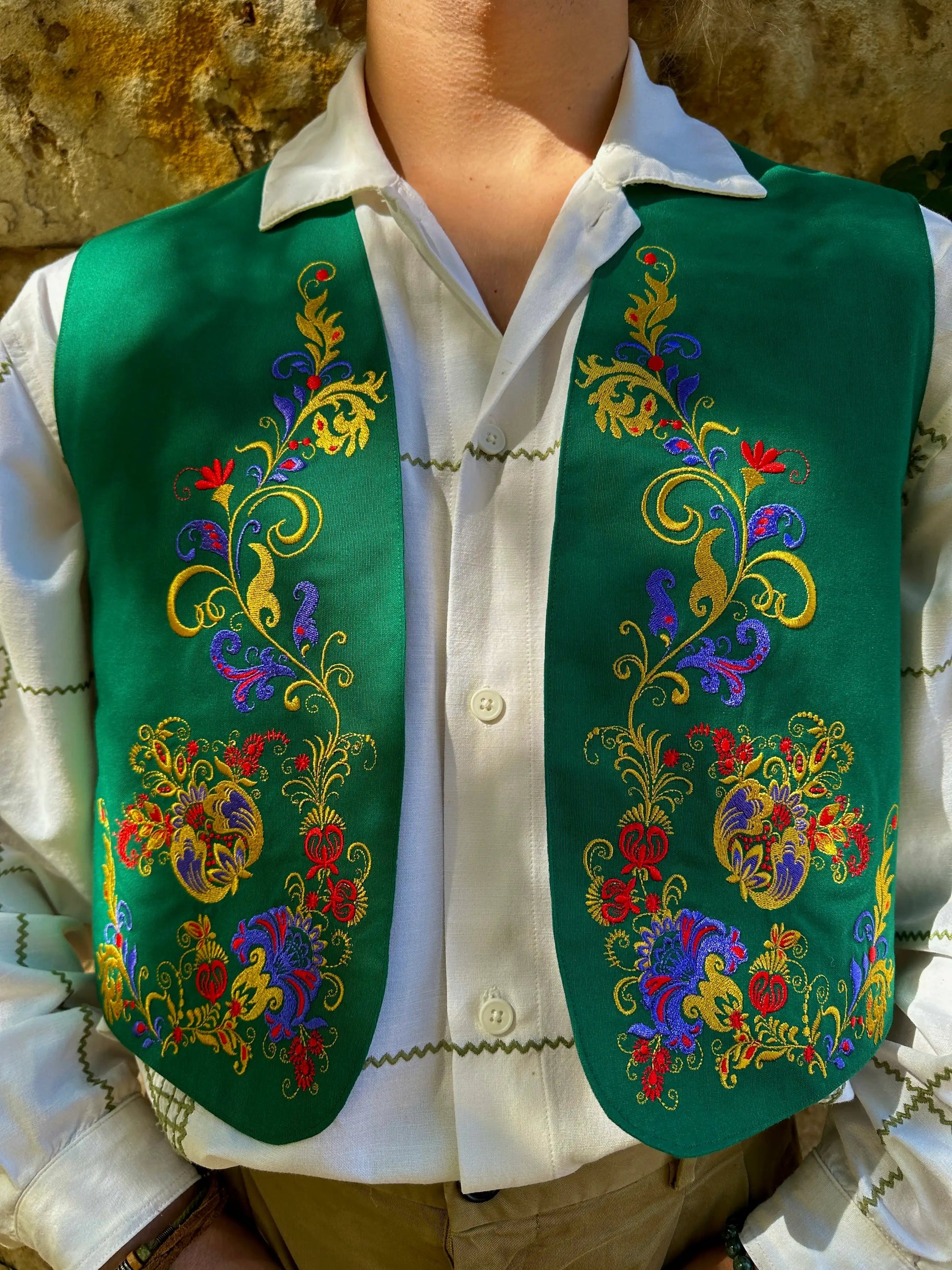 Person wearing a green embroidered vest over a white shirt with a stone wall background