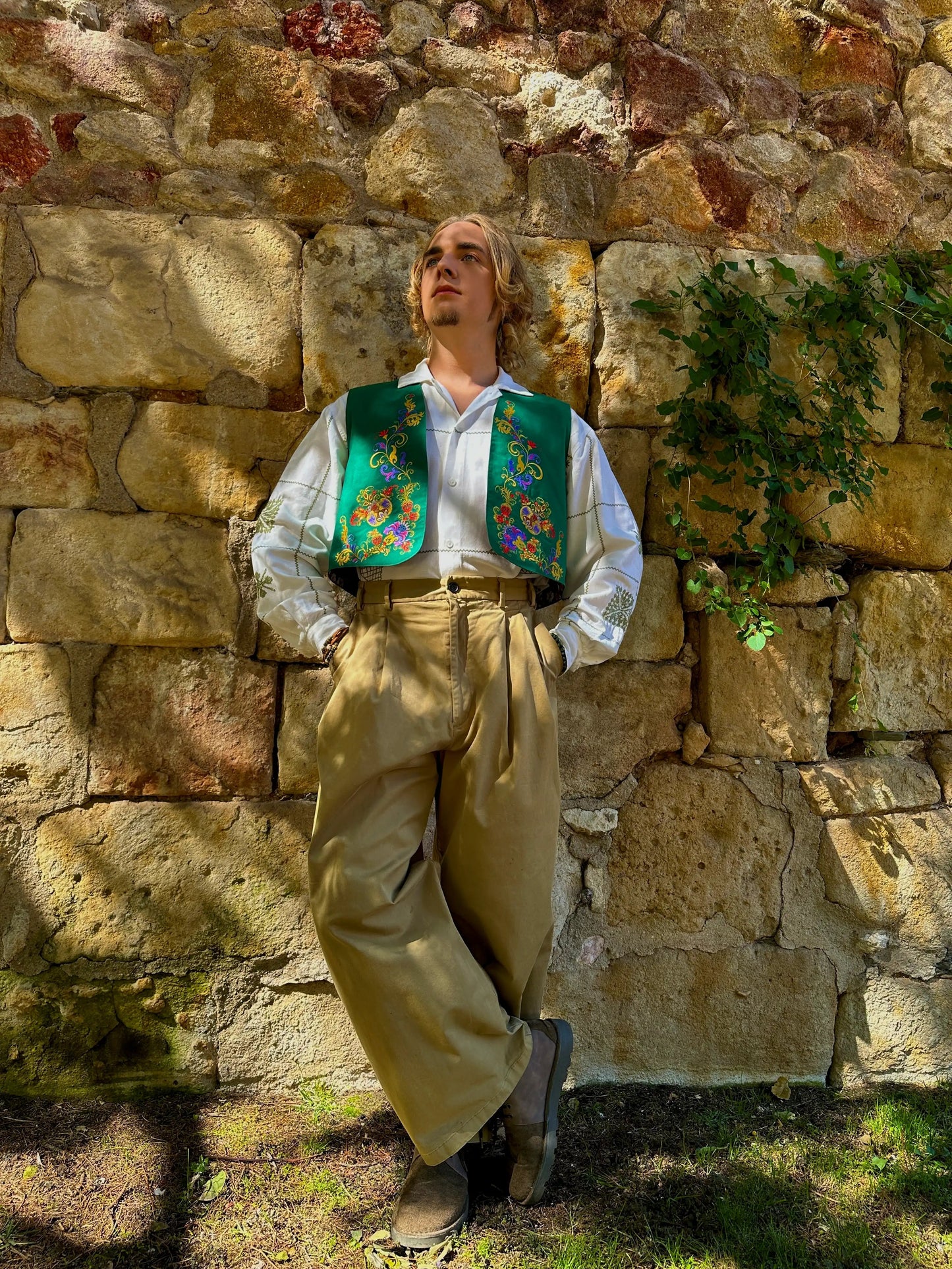 Person wearing a traditional outfit with a colorful vest against a stone wall.