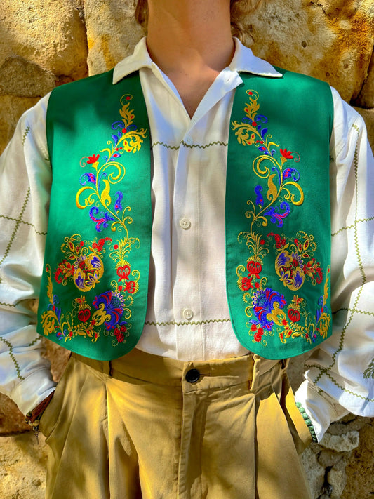 Mannequin wearing a white shirt and green embroidered vest with colorful floral patterns against a stone wall.