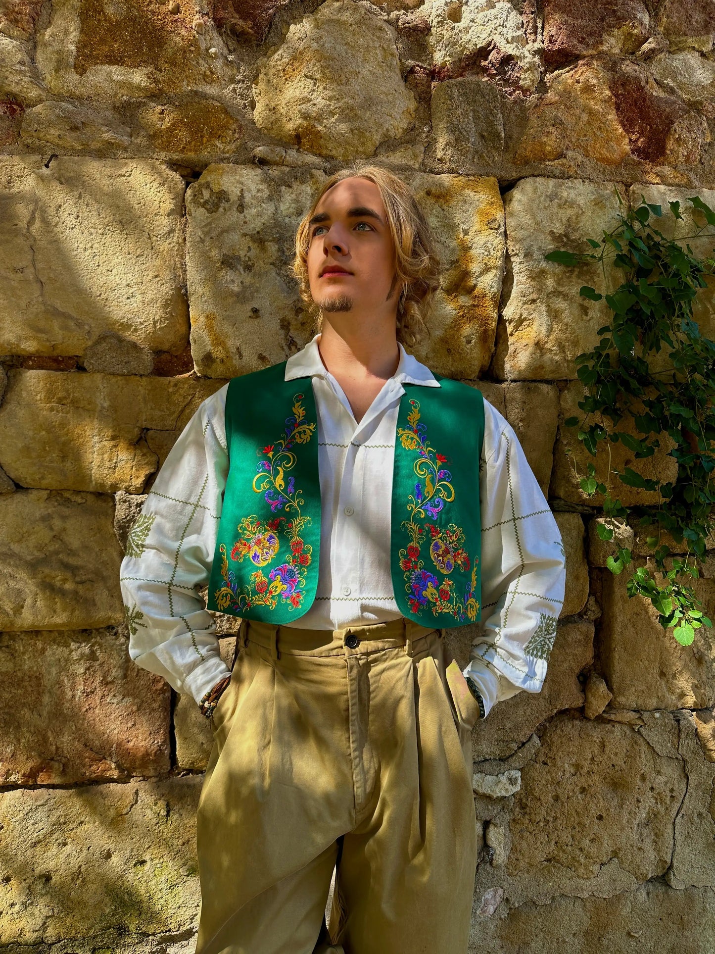 Person wearing a traditional outfit with a floral vest against a stone wall.