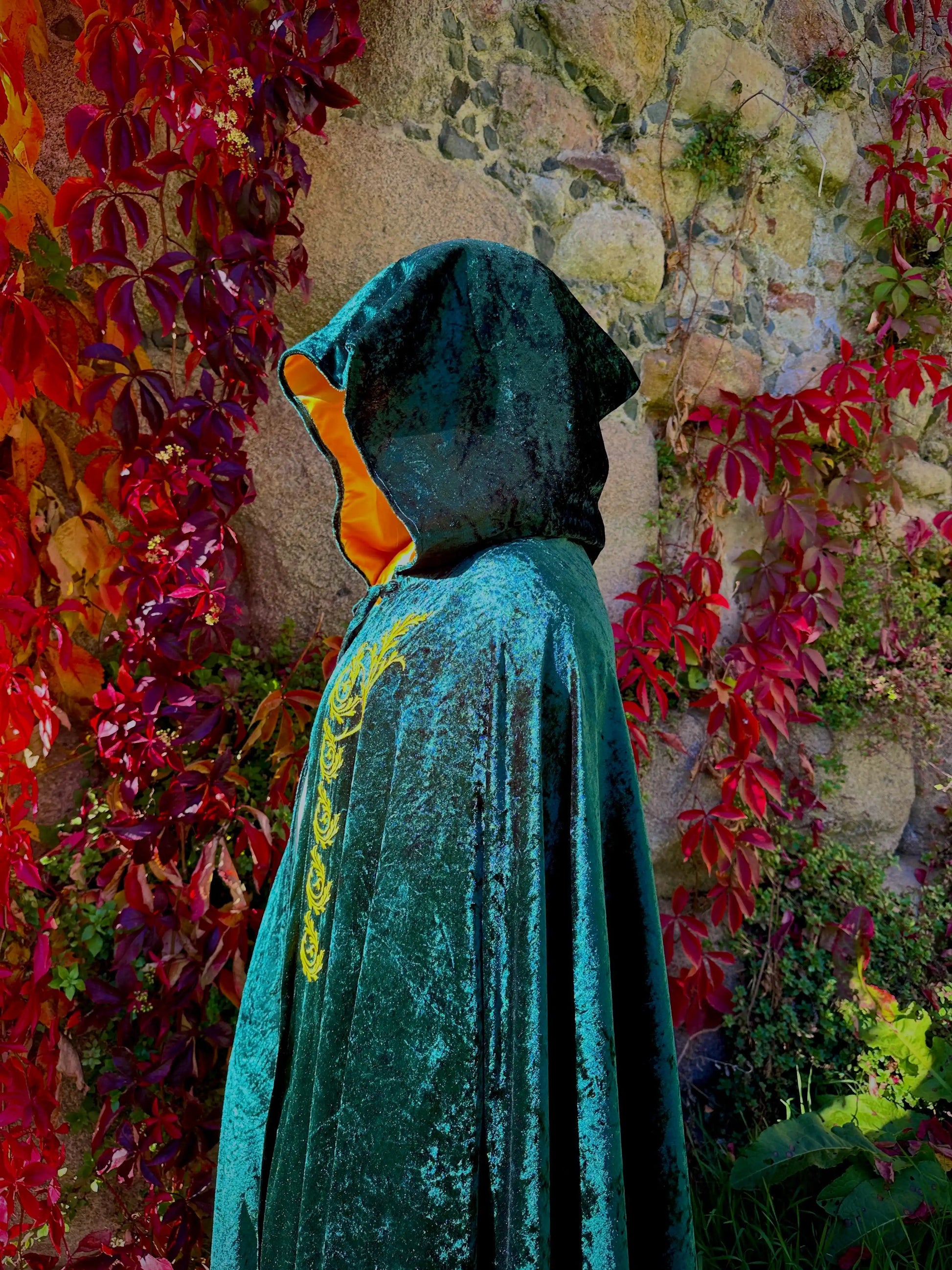 Person wearing a teal hooded cloak with gold embroidery against a colorful floral and stone wall background