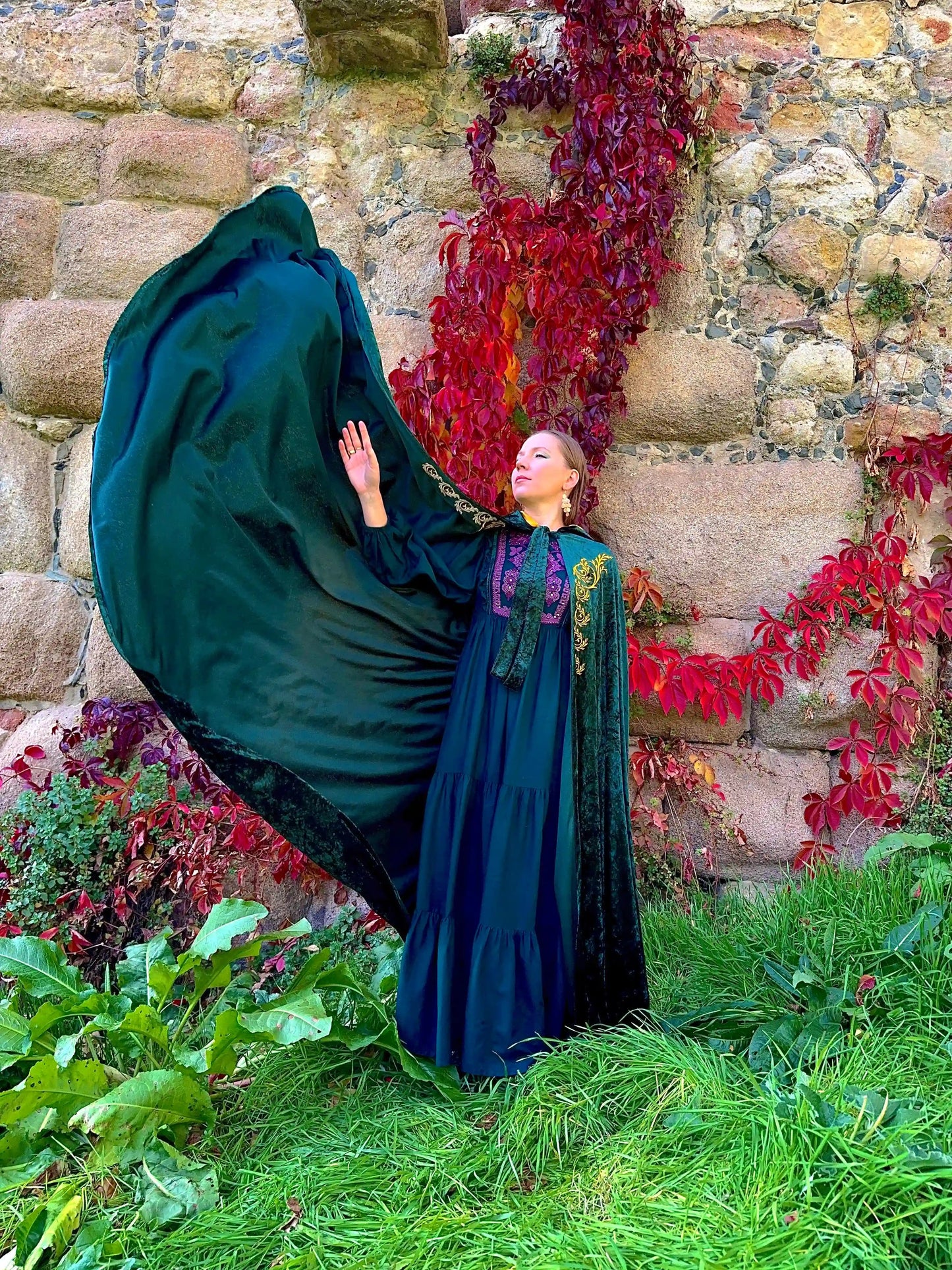 Person in a dark green cloak standing in front of a stone wall with red foliage