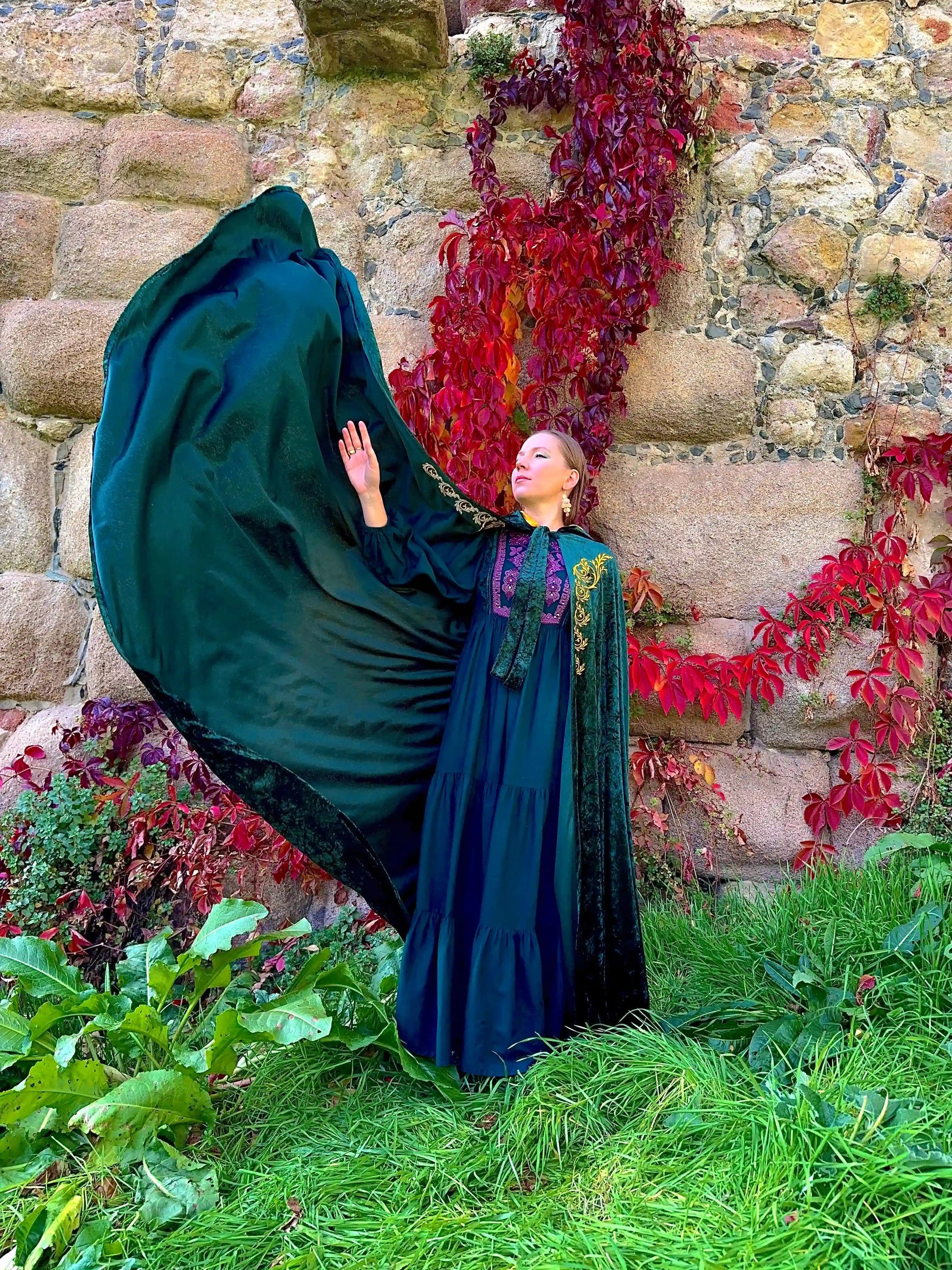 Person in a dark green cloak standing in front of a stone wall with red foliage