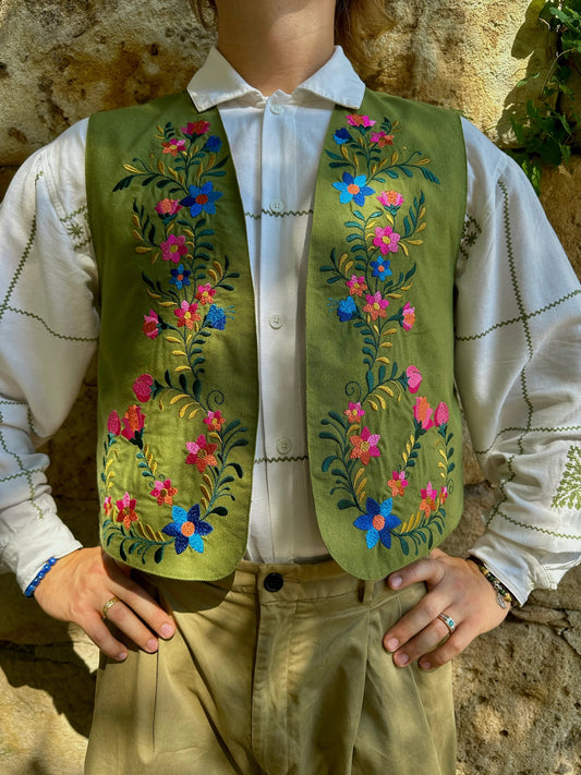 Person wearing a green embroidered vest over a white shirt against a stone wall.