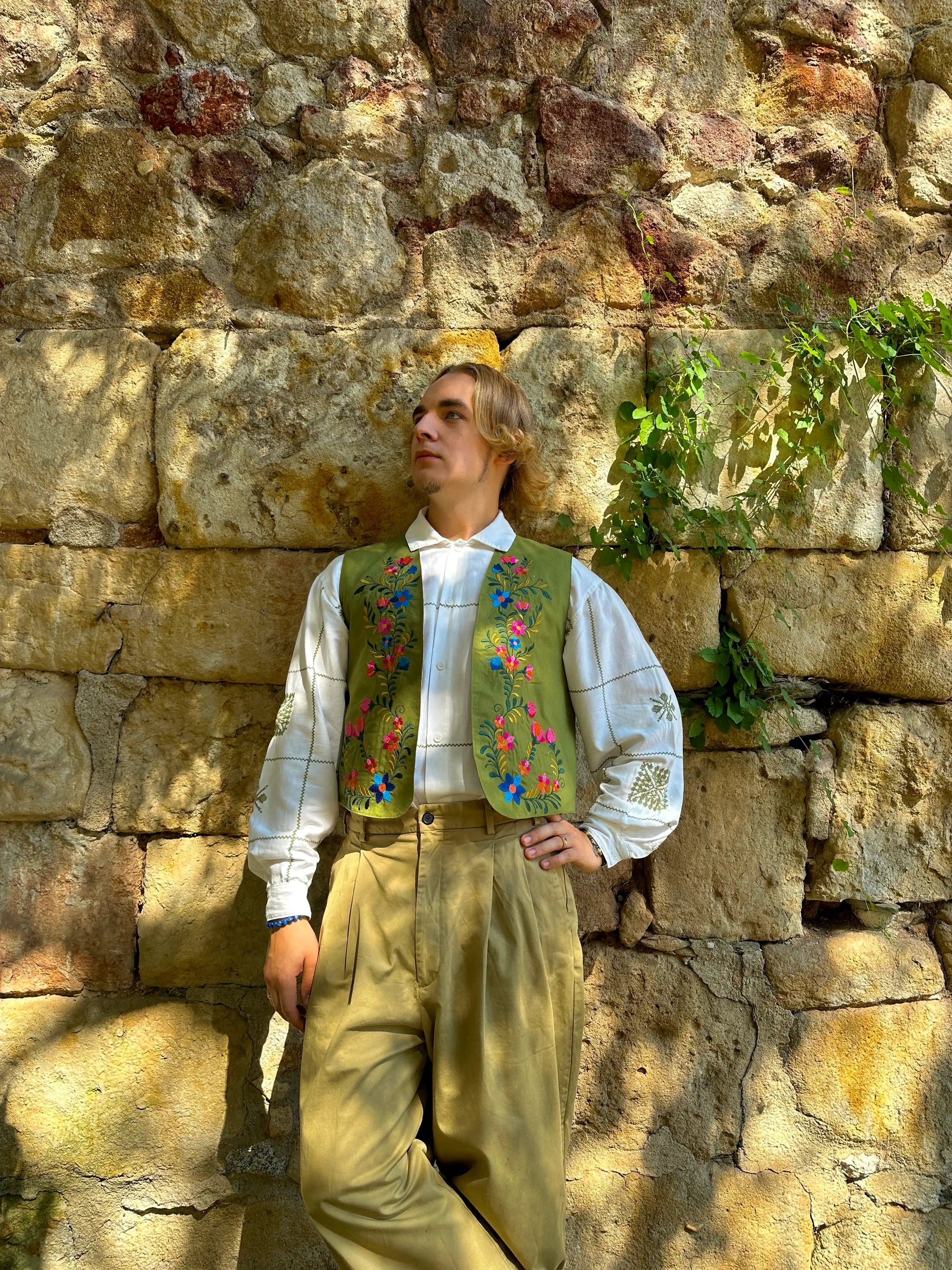Person wearing a traditional outfit with a colorful vest against a stone wall.
