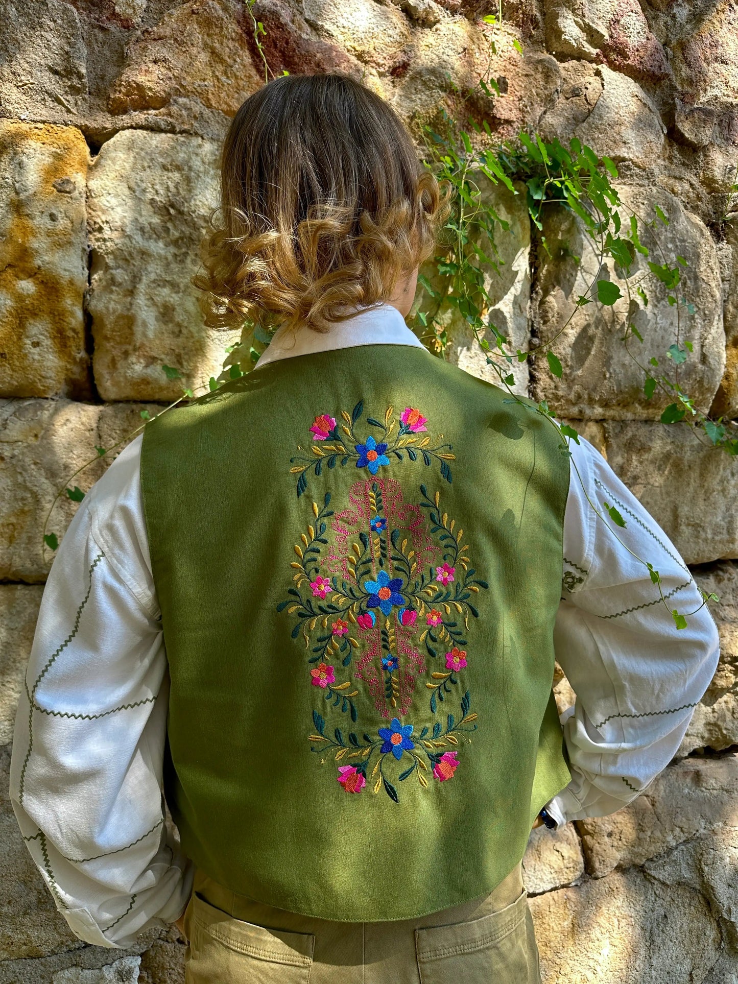 Person wearing a green embroidered vest standing against a stone wall.