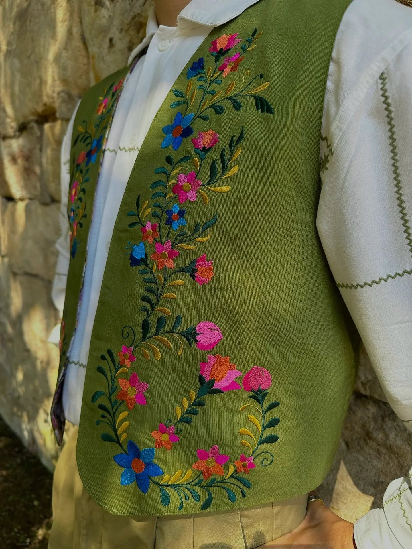 Green embroidered vest with colorful floral patterns worn over a white shirt against a stone wall.
