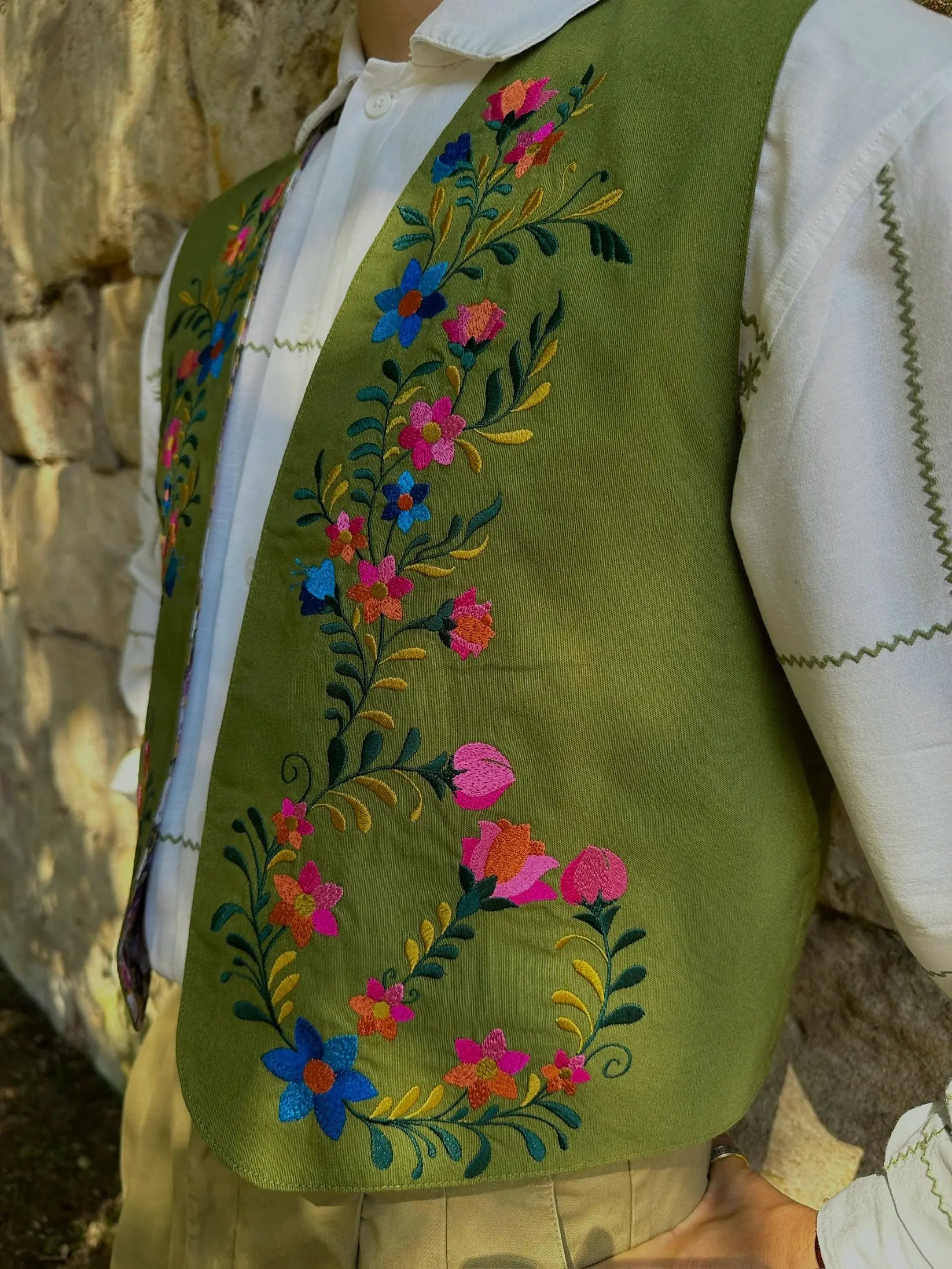 Green embroidered vest with colorful floral patterns worn over a white shirt against a stone wall.