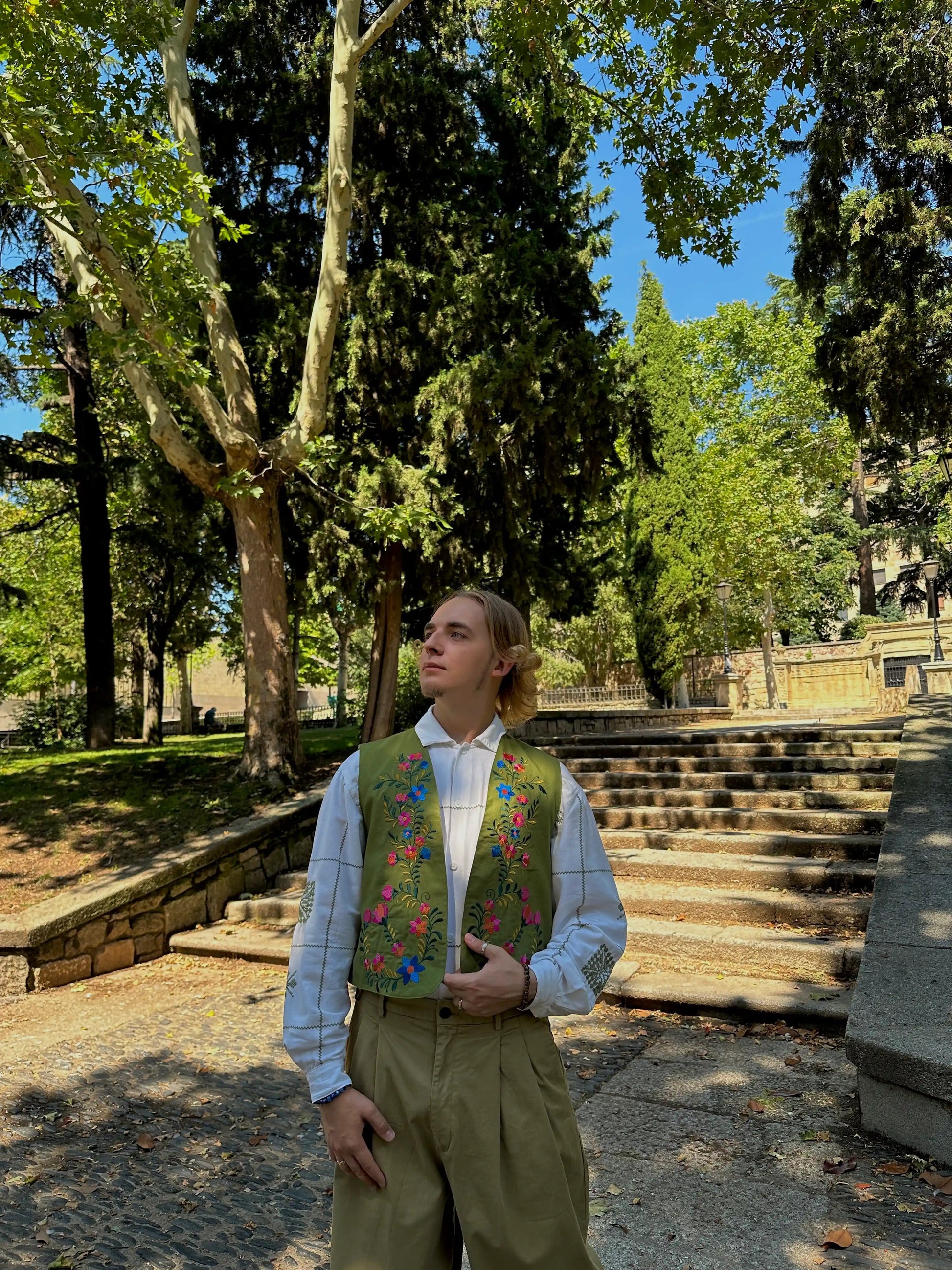 Person wearing a patterned vest standing in a park with trees and steps in the background