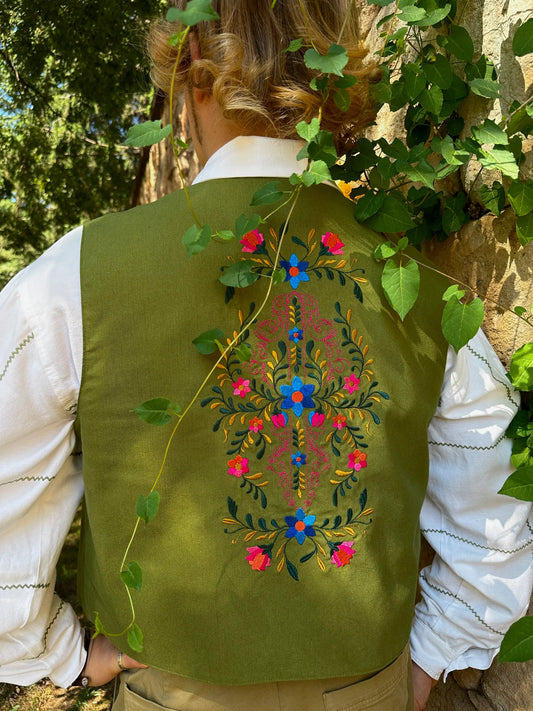 Person wearing a green vest with floral embroidery, standing among greenery.