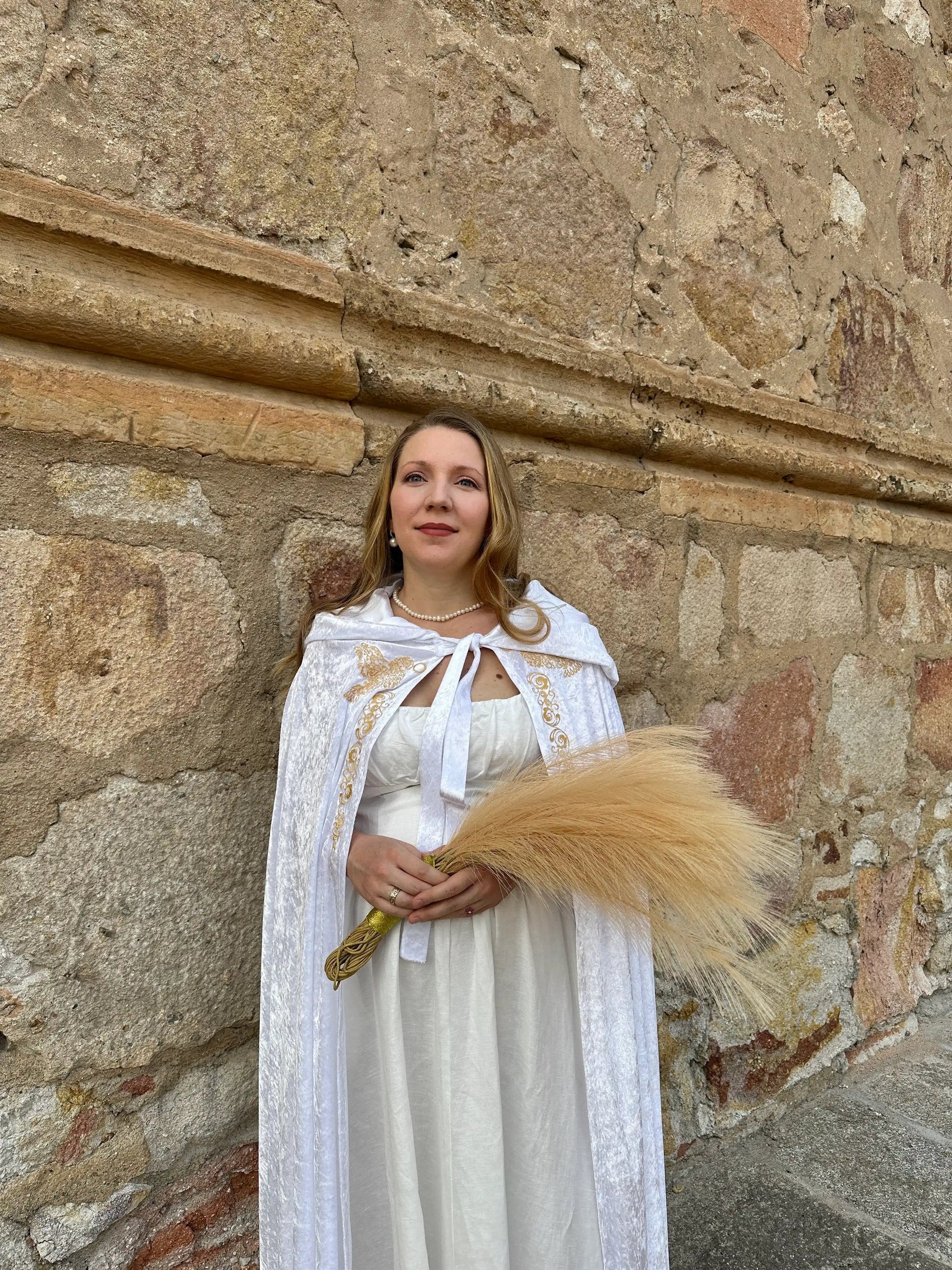 Woman in a white dress with a long, light-colored train standing against a stone wall.