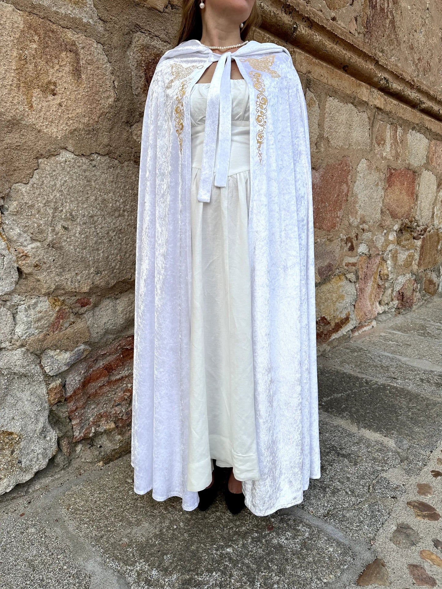 Person wearing a white lace dress with a cape against a stone wall.