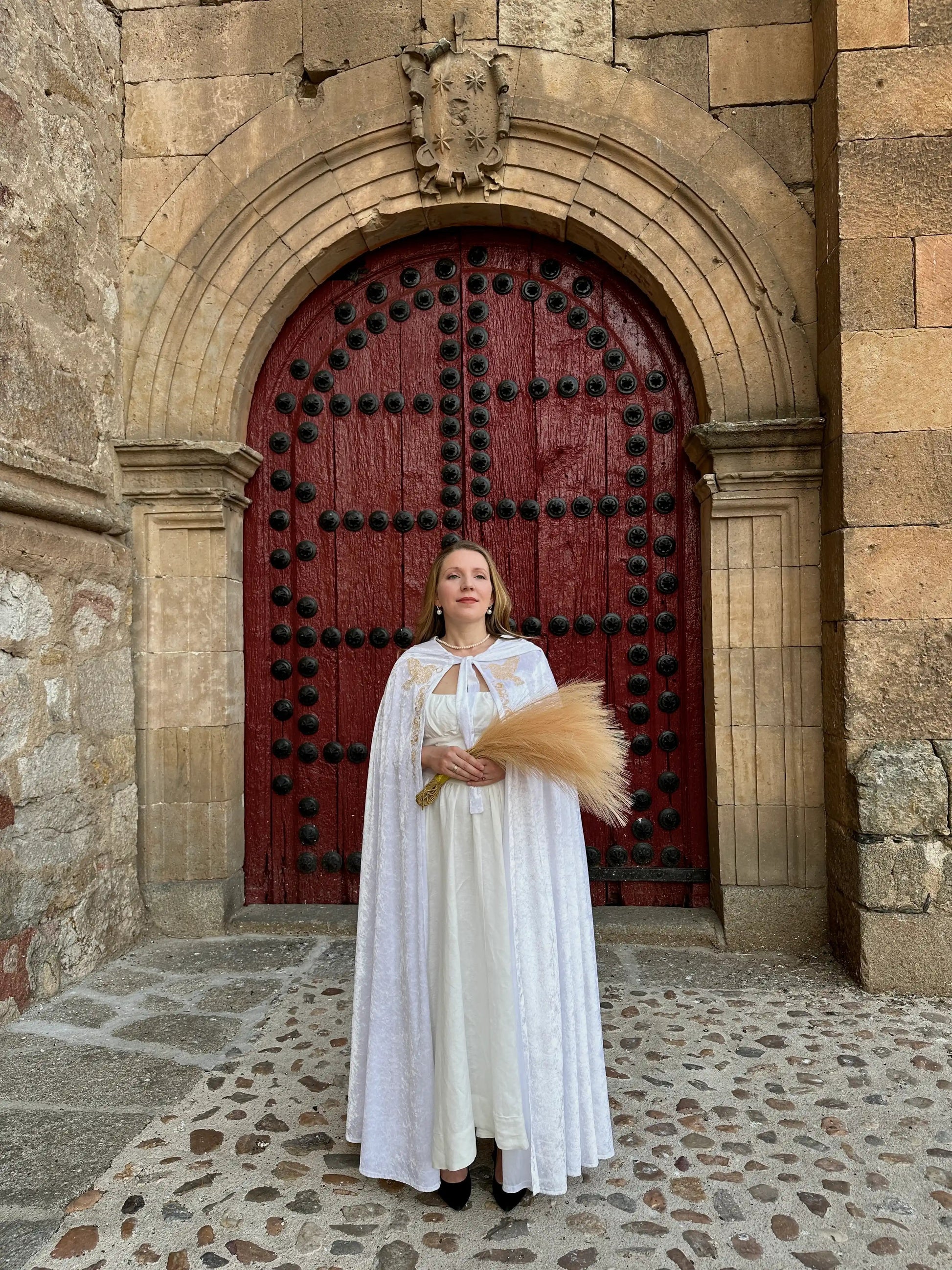 Woman in a white dress standing in front of a large red door with stone architecture.