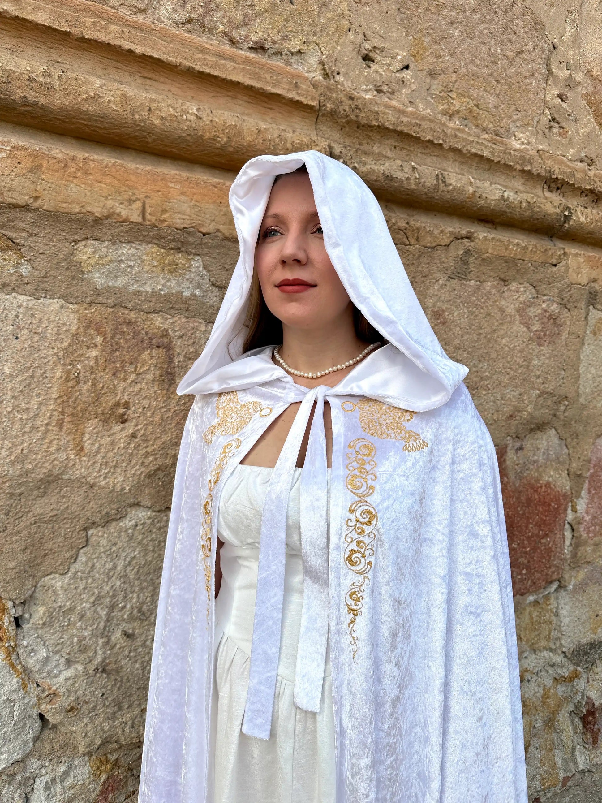 Woman wearing a white cape with gold embroidery against a stone wall.