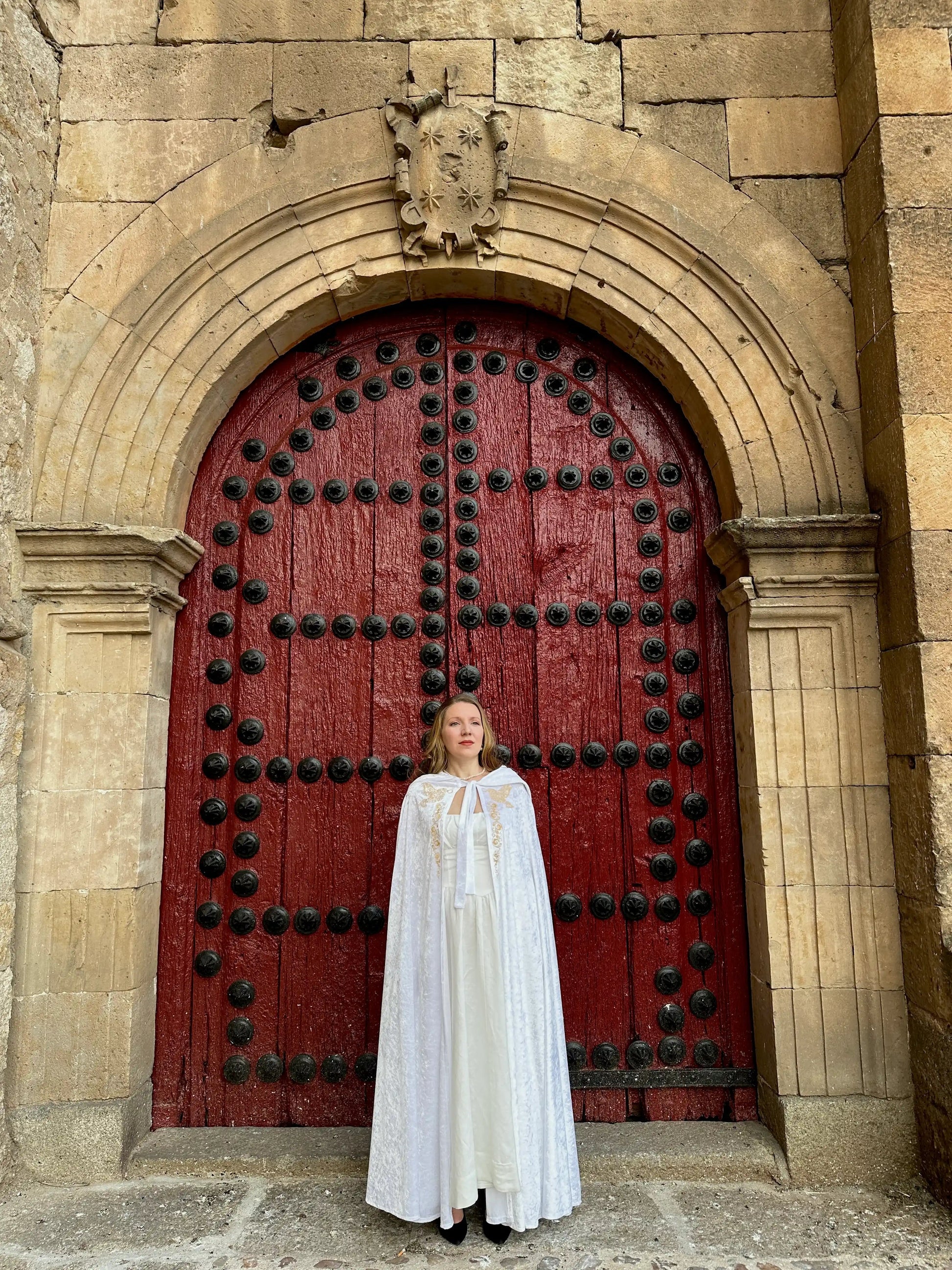 Person in a white cloak standing in front of a large red door with black metalwork on a stone building.