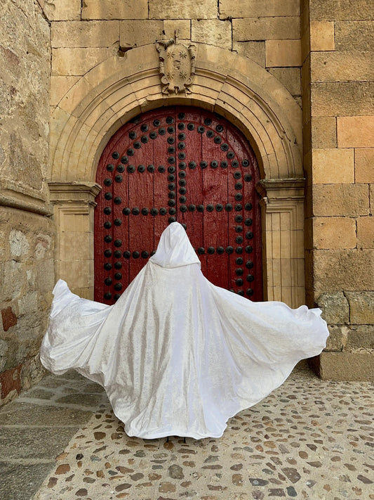 Person in a white hooded garment standing in front of a red door on a stone pathway.