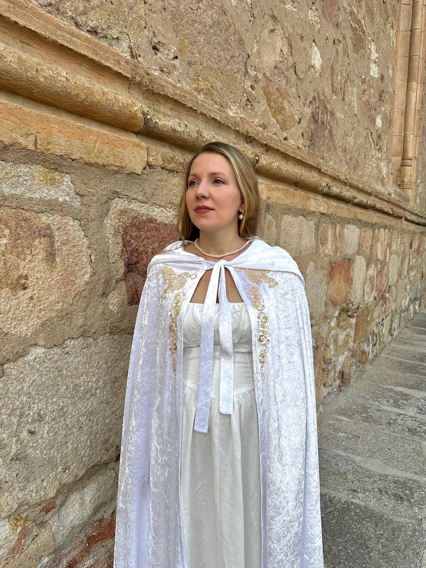 Woman in a white dress with a cape standing against a stone wall.