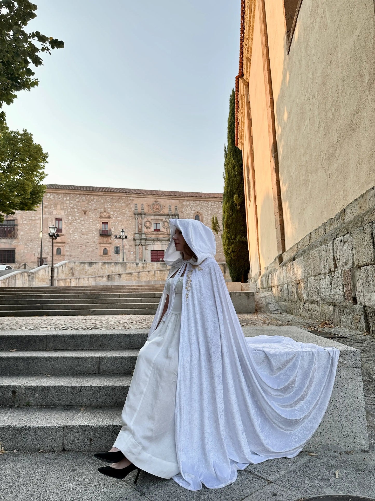 Person in a white wedding dress with a hooded cape standing on stone steps outdoors.