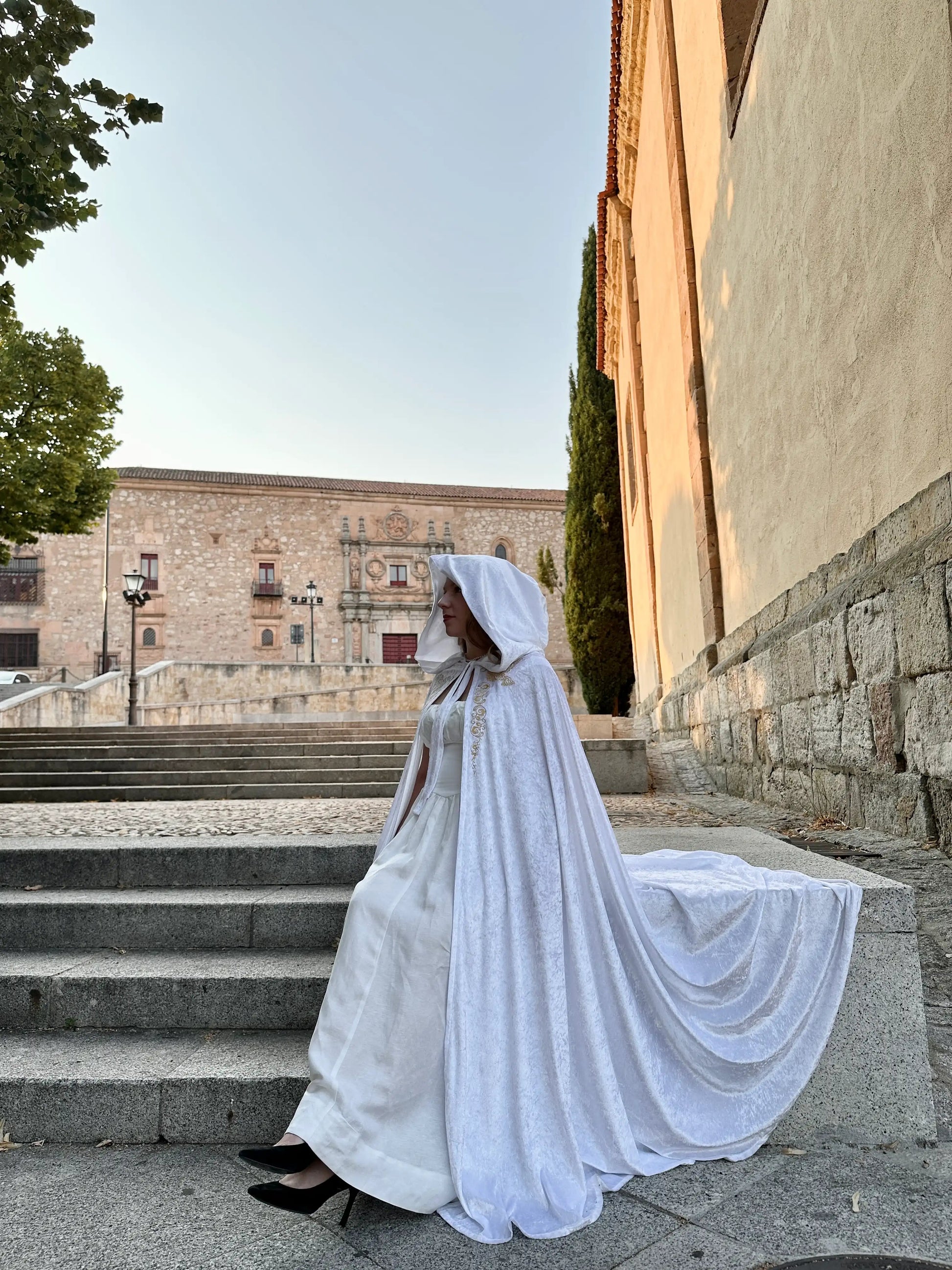 Person in a white wedding dress with a hooded cape standing on stone steps outdoors.